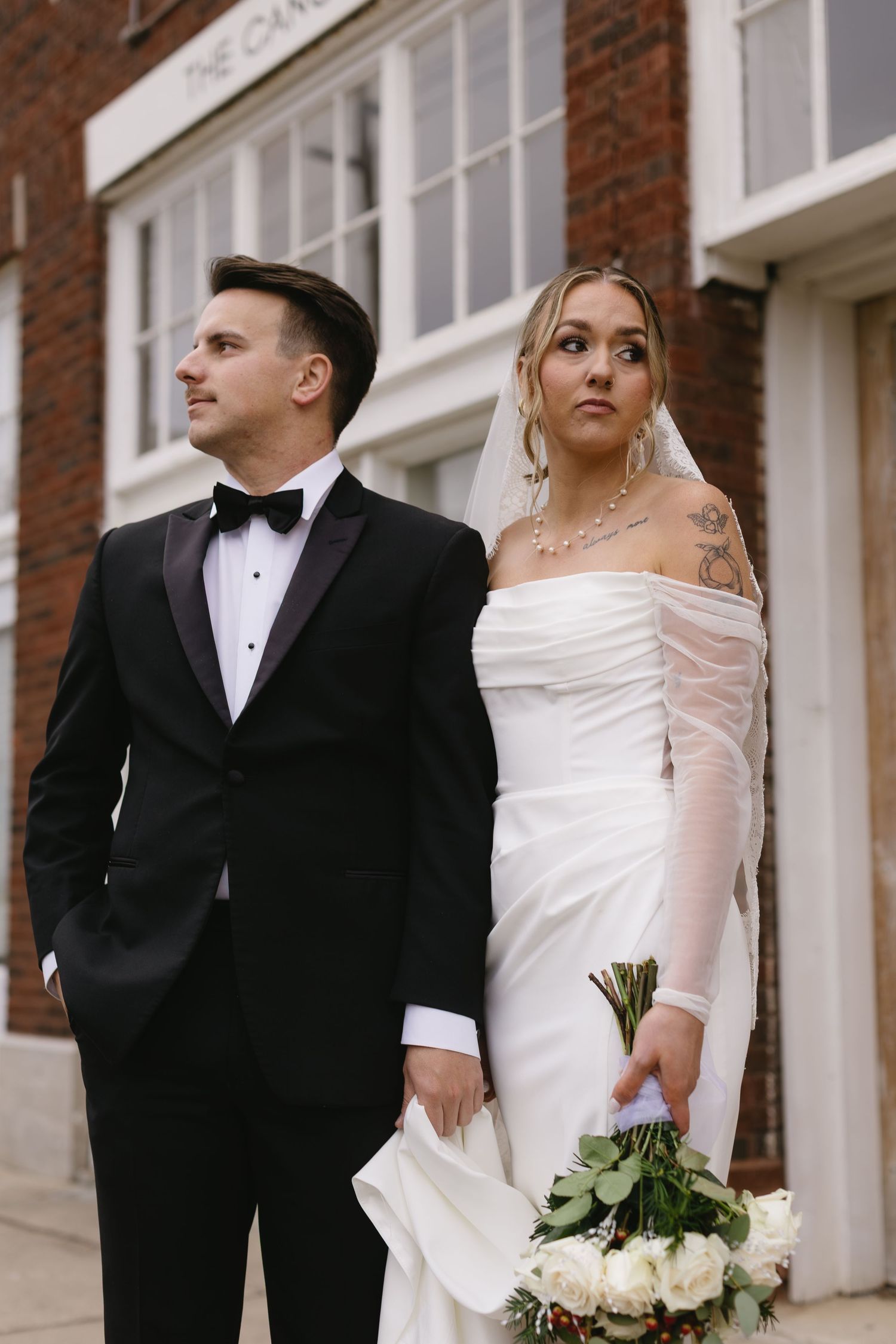 A couple in wedding attire poses outside a brick building with white trim and windows.