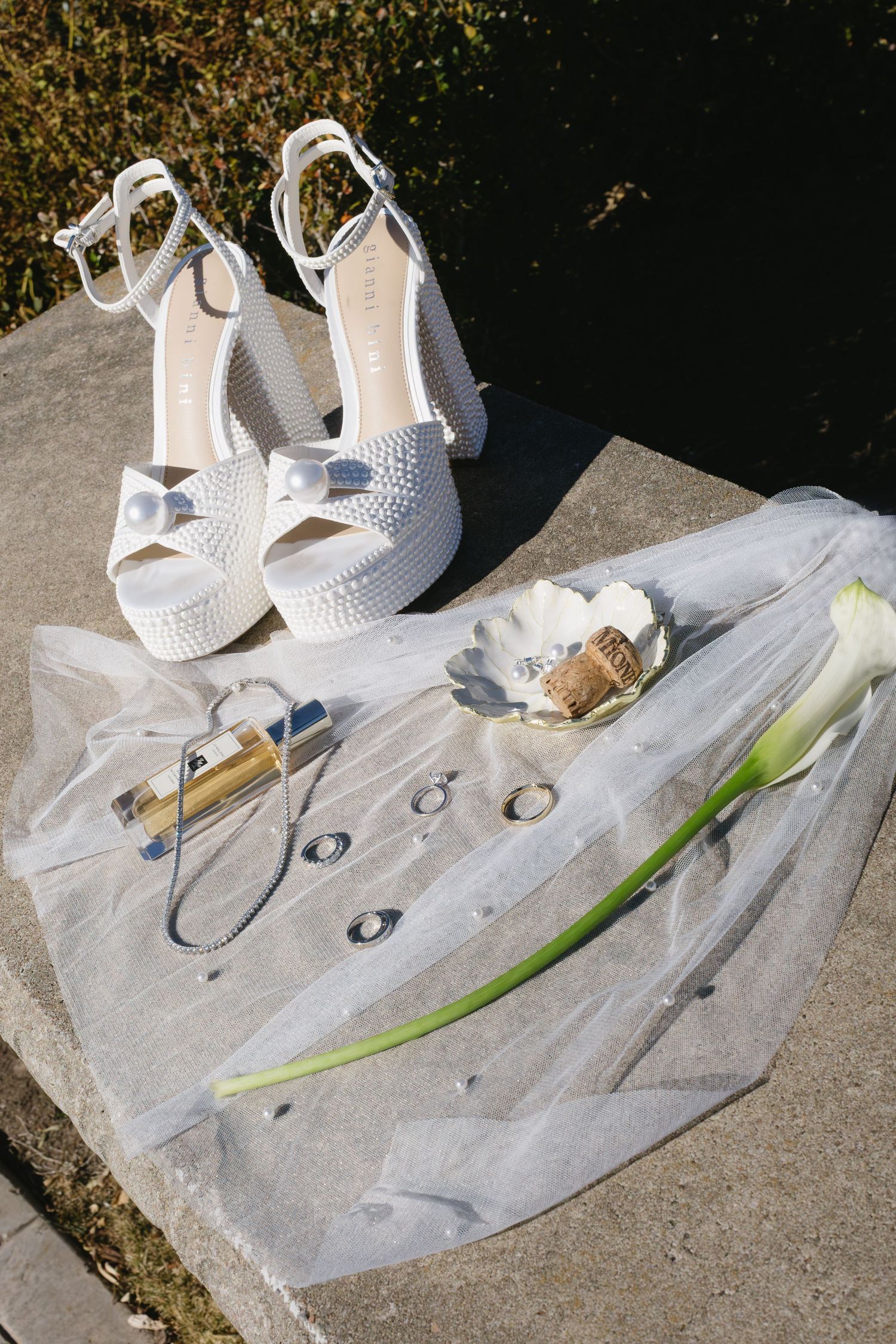 White bridal shoes, wedding rings, and a single flower stem arranged on a white veil against concrete background.
