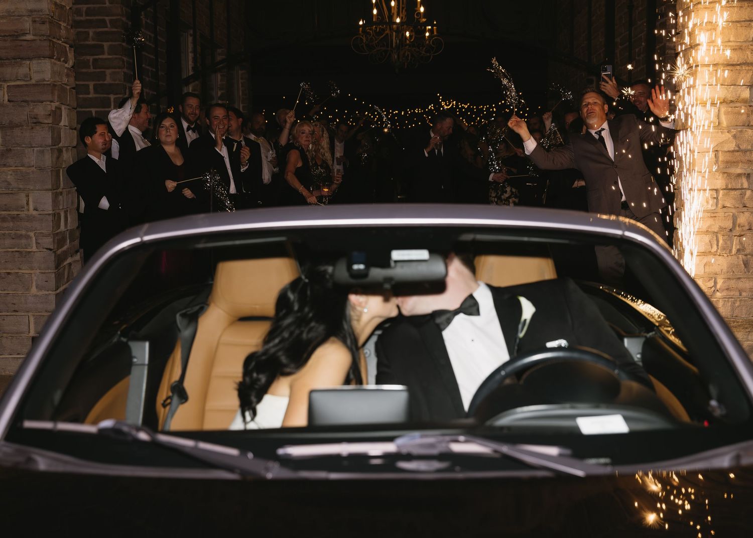 Wedding guests gathered around parked cars at night with string lights in the background.