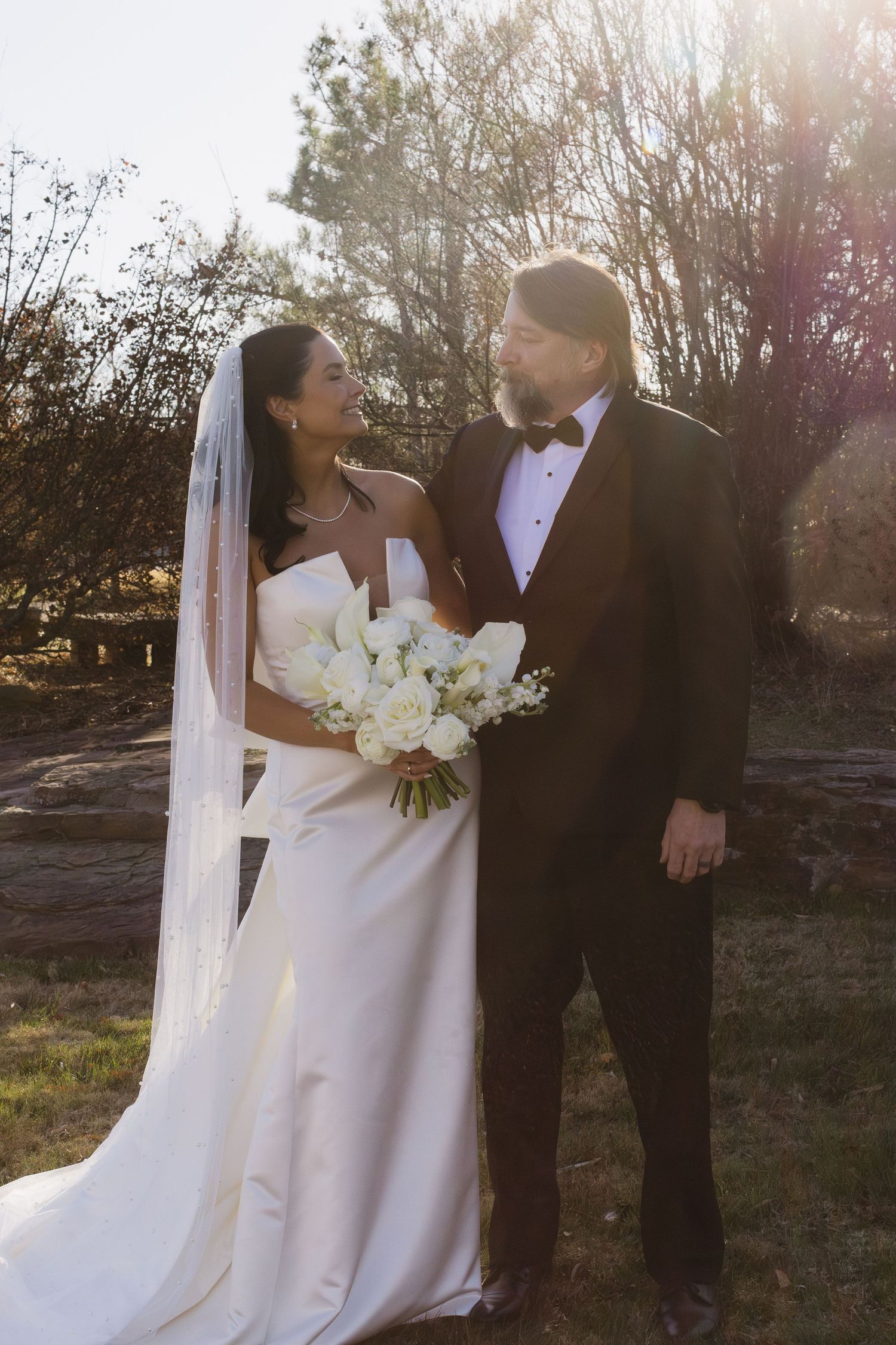 Romantic wedding couple poses in outdoor setting with sunlight filtering through trees.