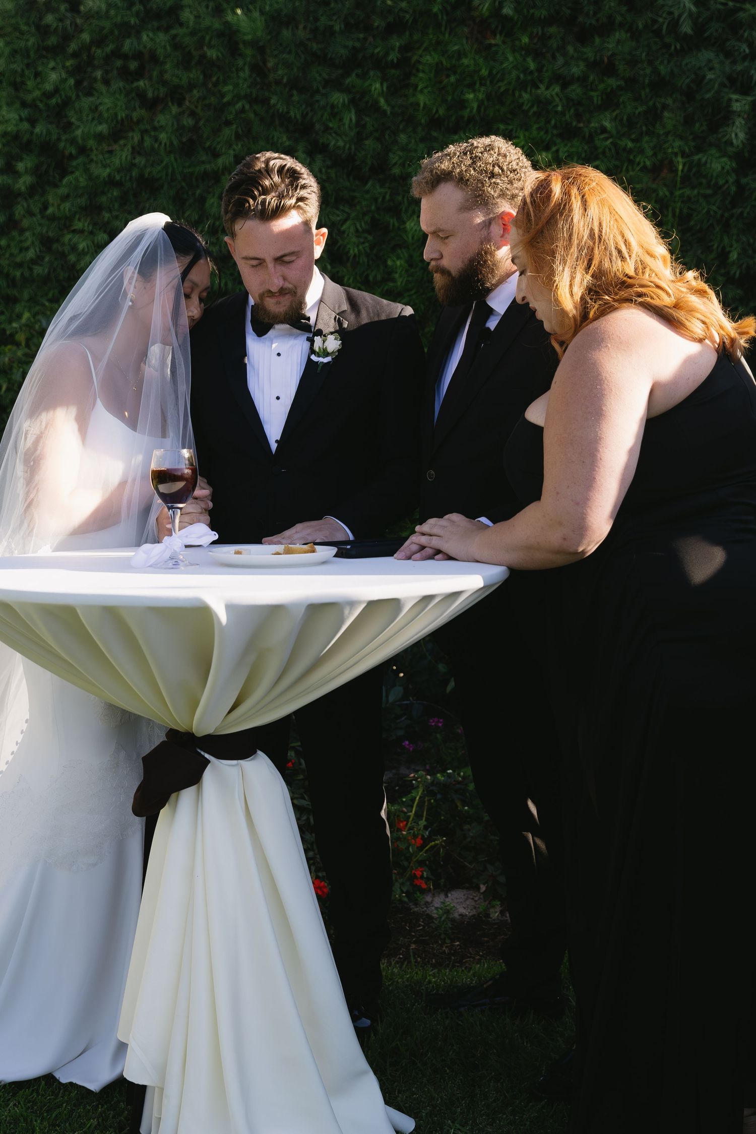 Wedding party members in formal attire gather around a white decorative element outdoors.