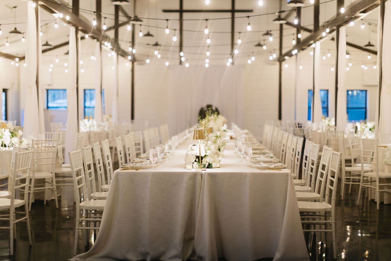 A bright white wedding reception hall with elegant table settings and hanging lights creating a romantic atmosphere.