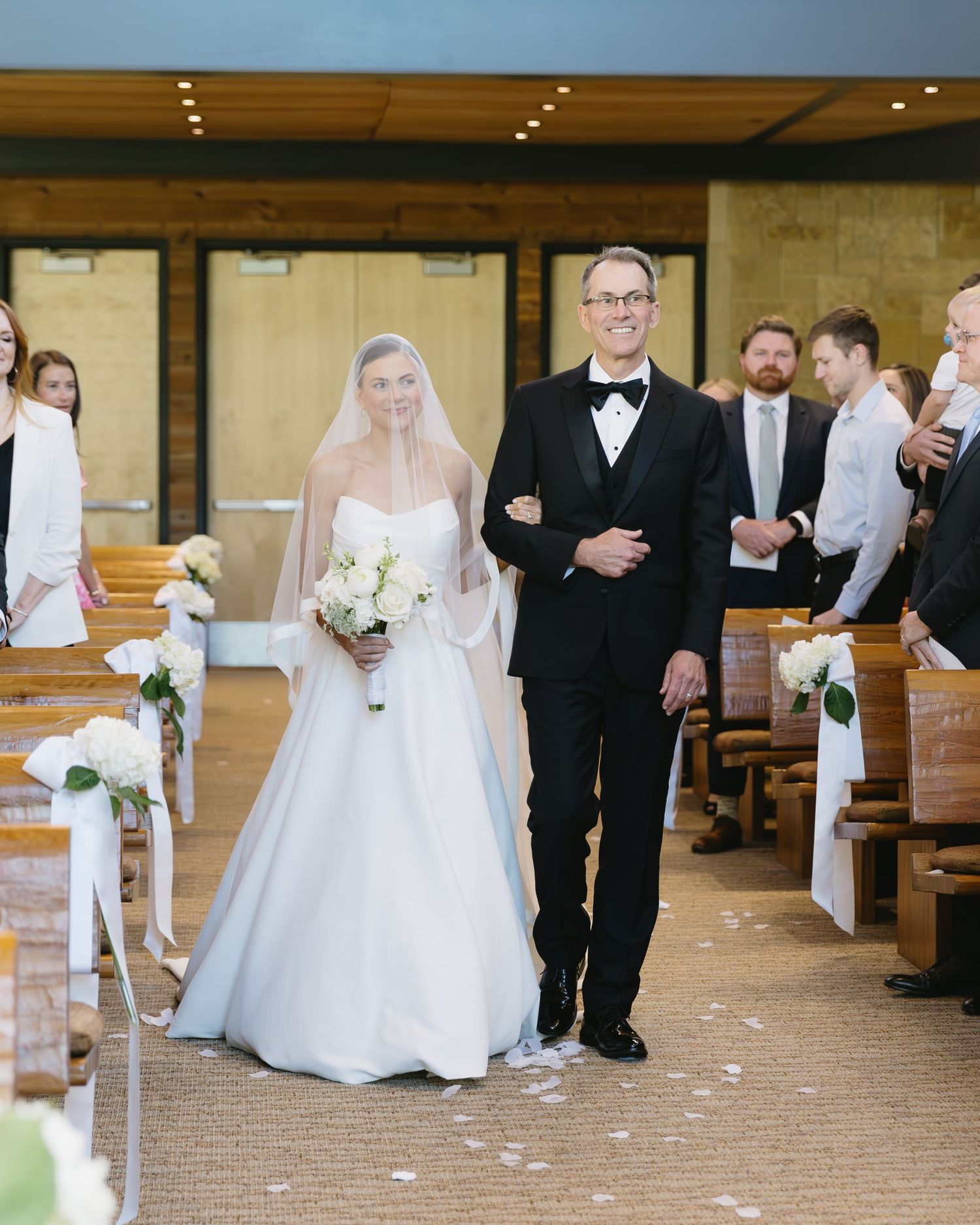 A traditional wedding ceremony procession in a wooden chapel with floral aisle decorations.