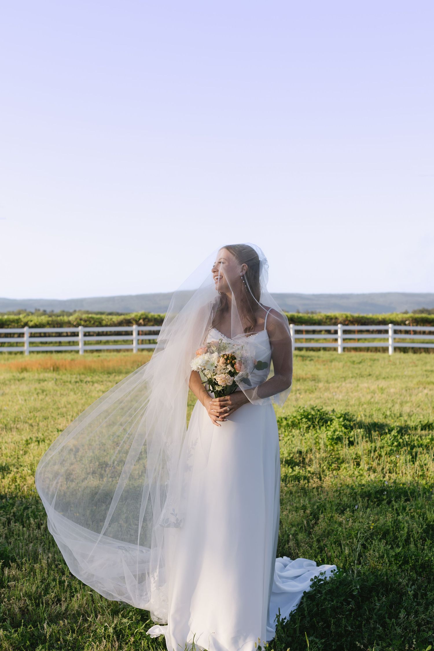 Bride in flowing white dress poses in an open field with white fence and countryside views.