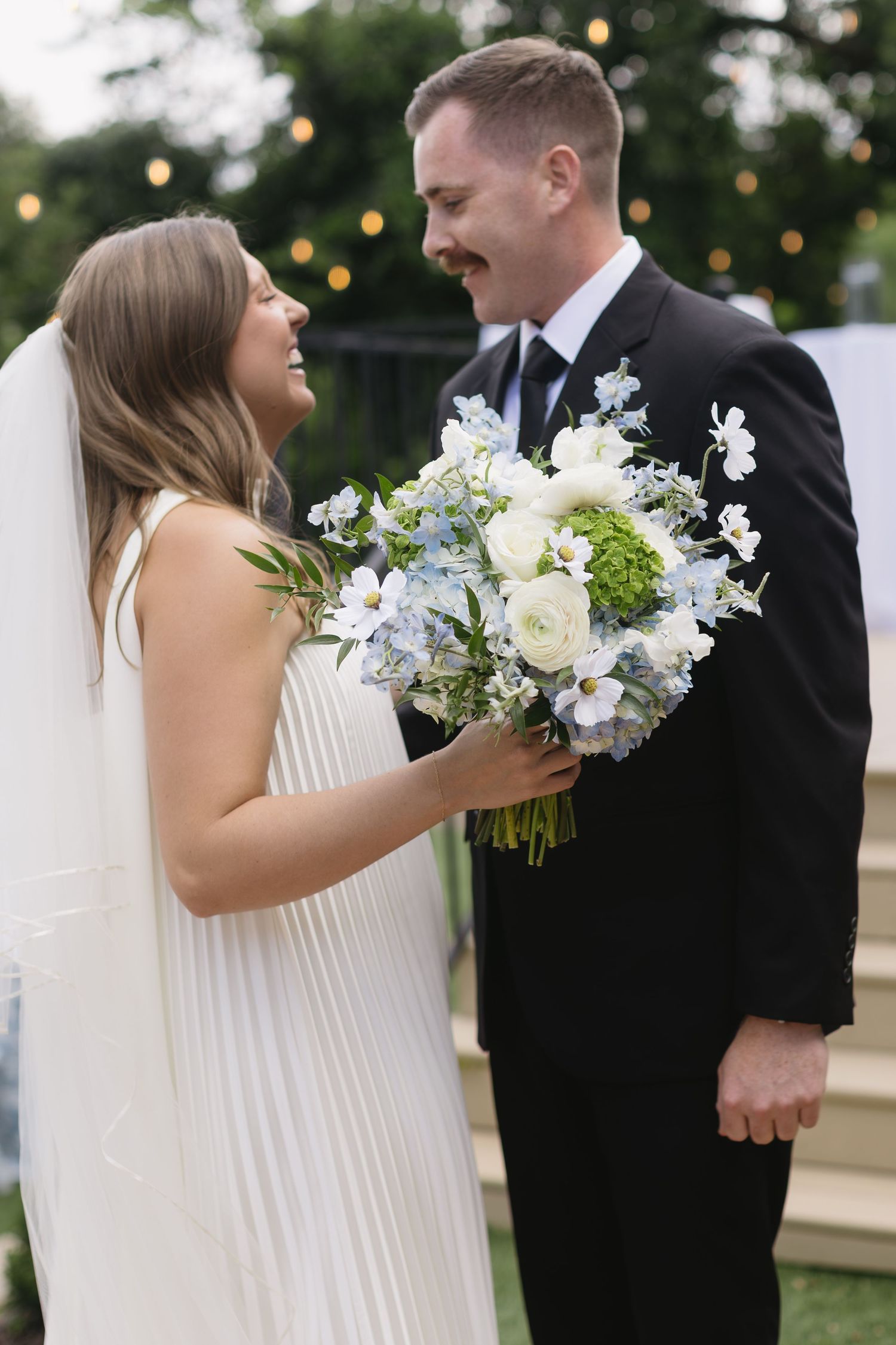 Wedding couple sharing intimate moments outdoors with white floral bouquet and string lights in background.