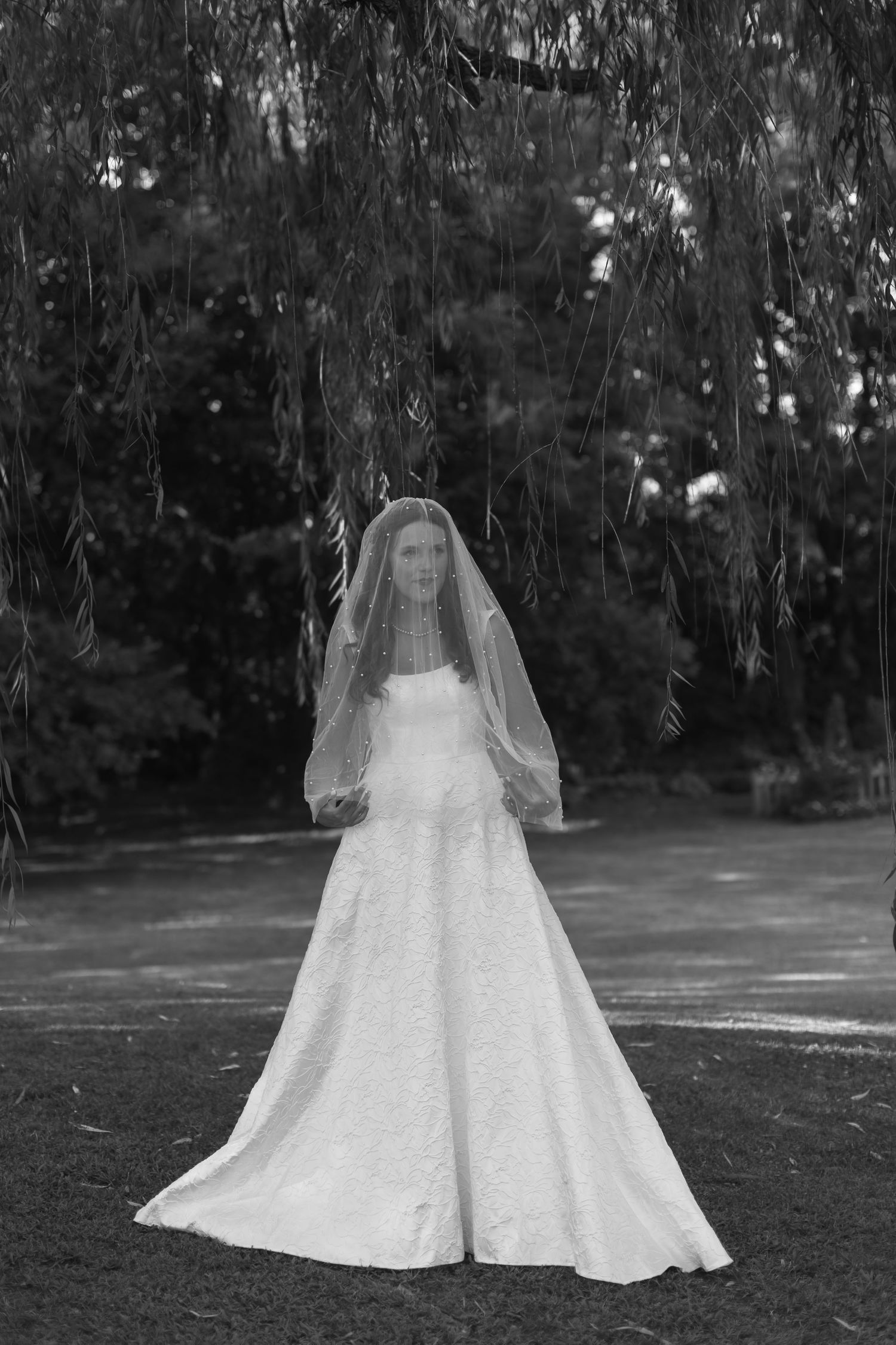 A black and white portrait of a bride in a flowing white wedding dress and veil standing outdoors under trees.