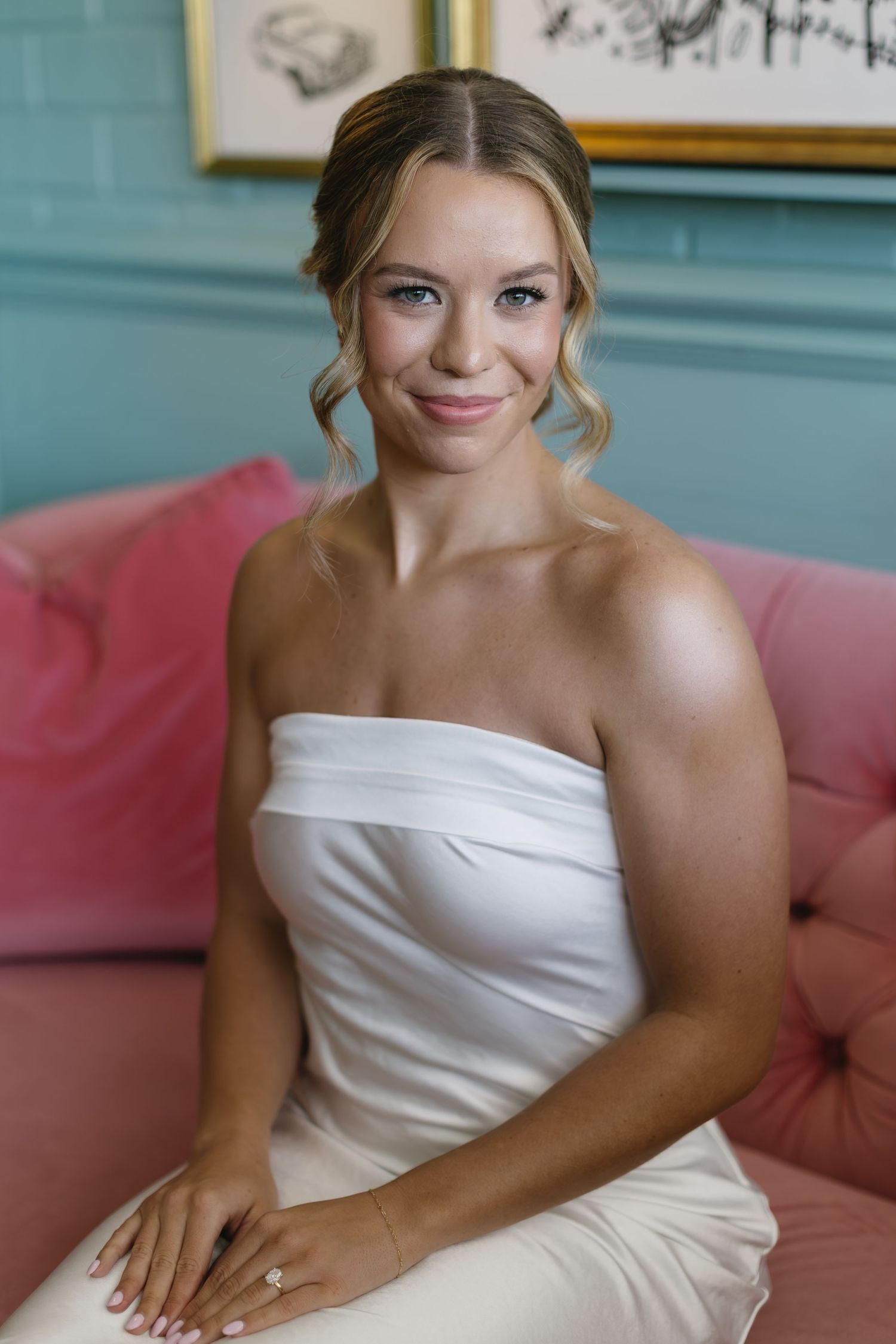 Elegant bride in white strapless dress sits on pink couch during pre-wedding preparations.