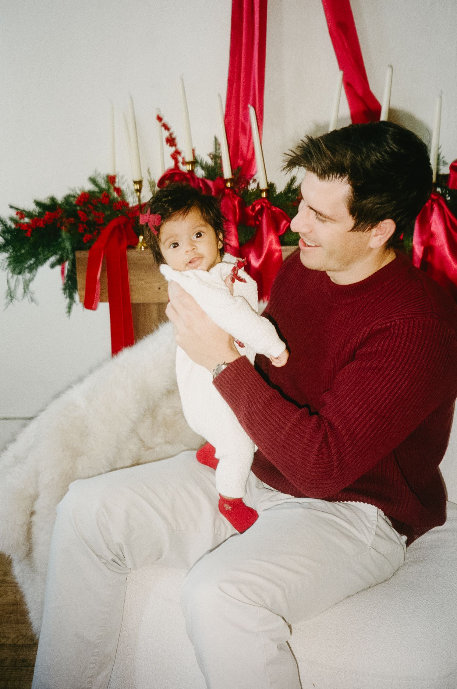Baby in white outfit being held near Christmas decorations with red ribbons and greenery.