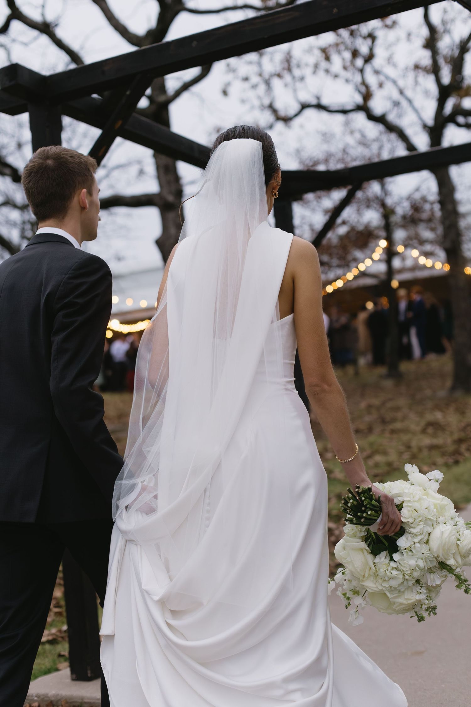 A bride in a flowing white dress and groom in black suit walk together outdoors with string lights visible.