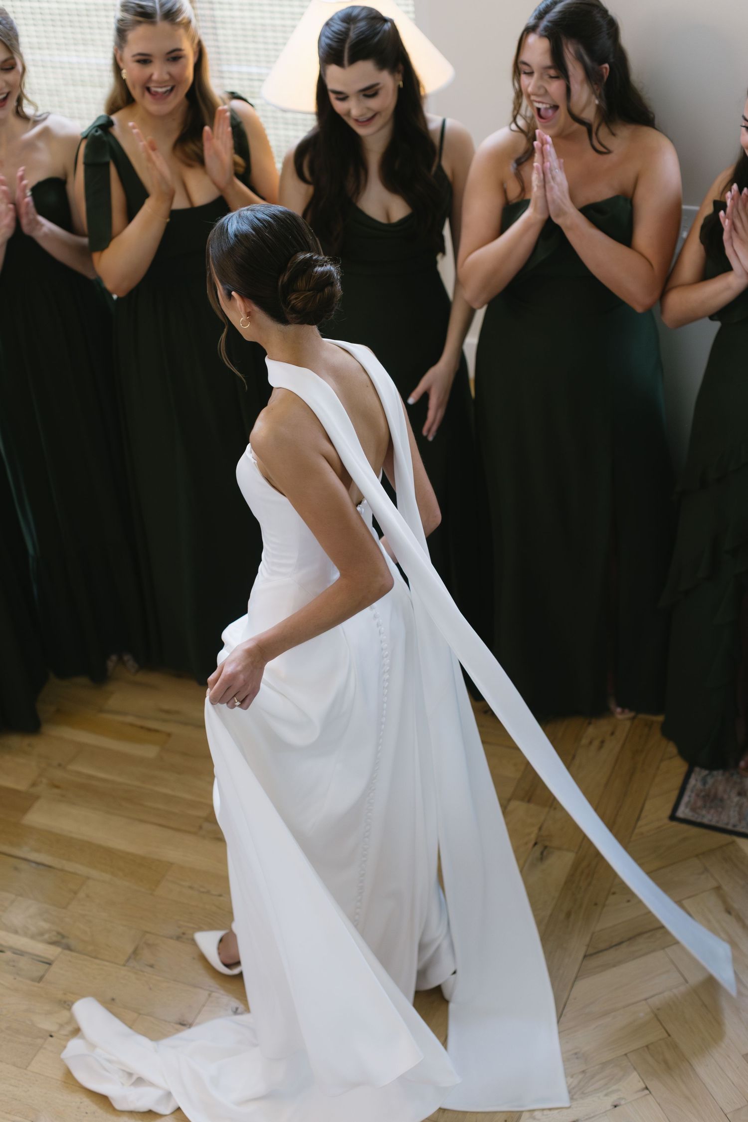 Bridesmaids in black dresses react joyfully as bride twirls in elegant white wedding gown during pre-ceremony preparations.