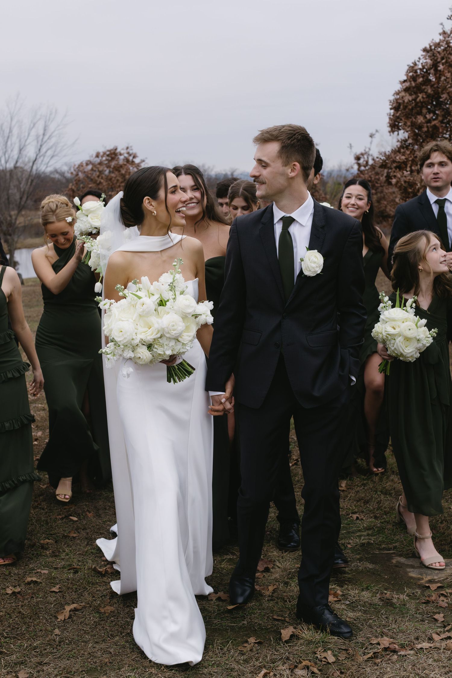 Wedding party gathered outdoors in autumn with bridesmaids in black dresses and white bouquets.