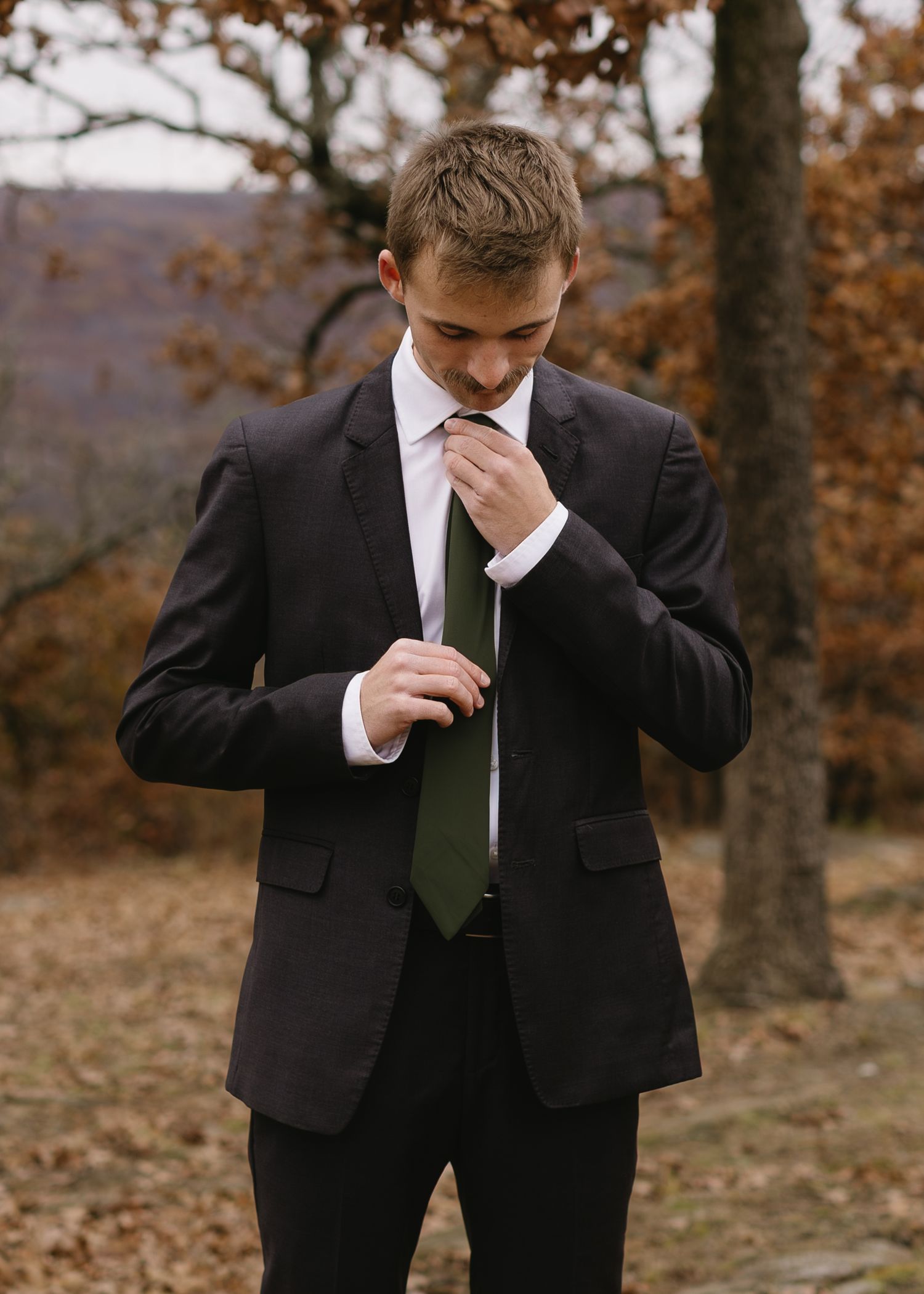 A person in a black suit and green tie adjusts their necktie while standing in an autumn forest setting.