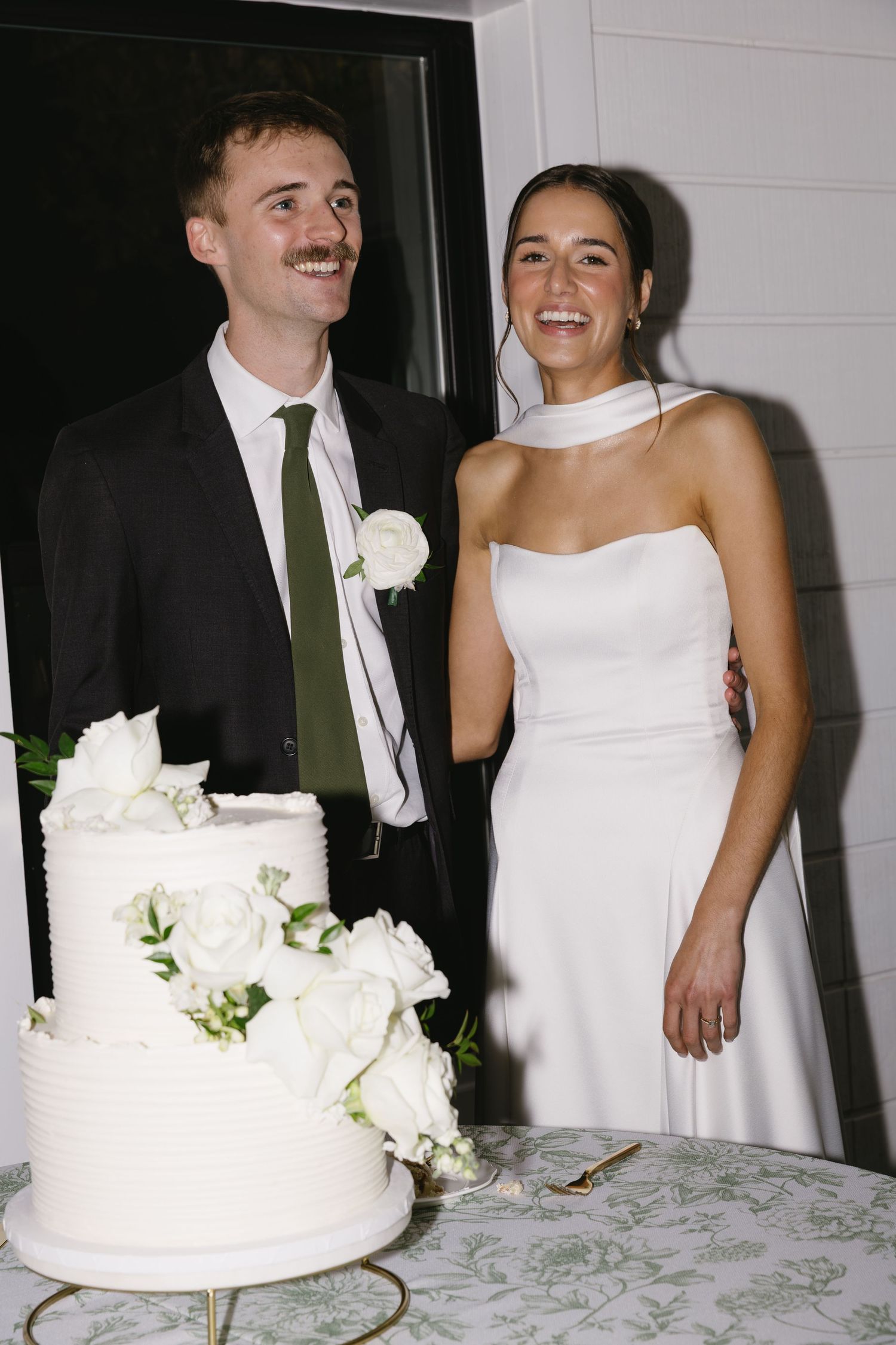 Newlyweds stand beside their elegant white wedding cake decorated with roses.
