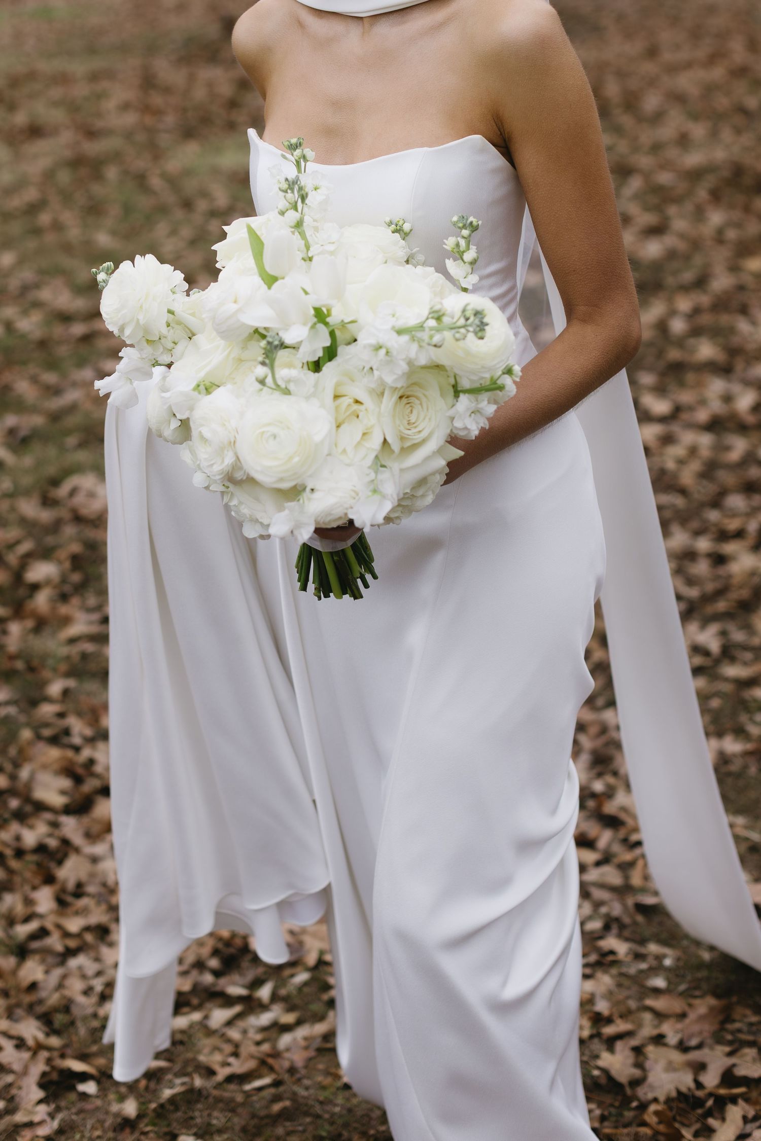 White wedding dress with white floral bouquet against autumn leaves background.