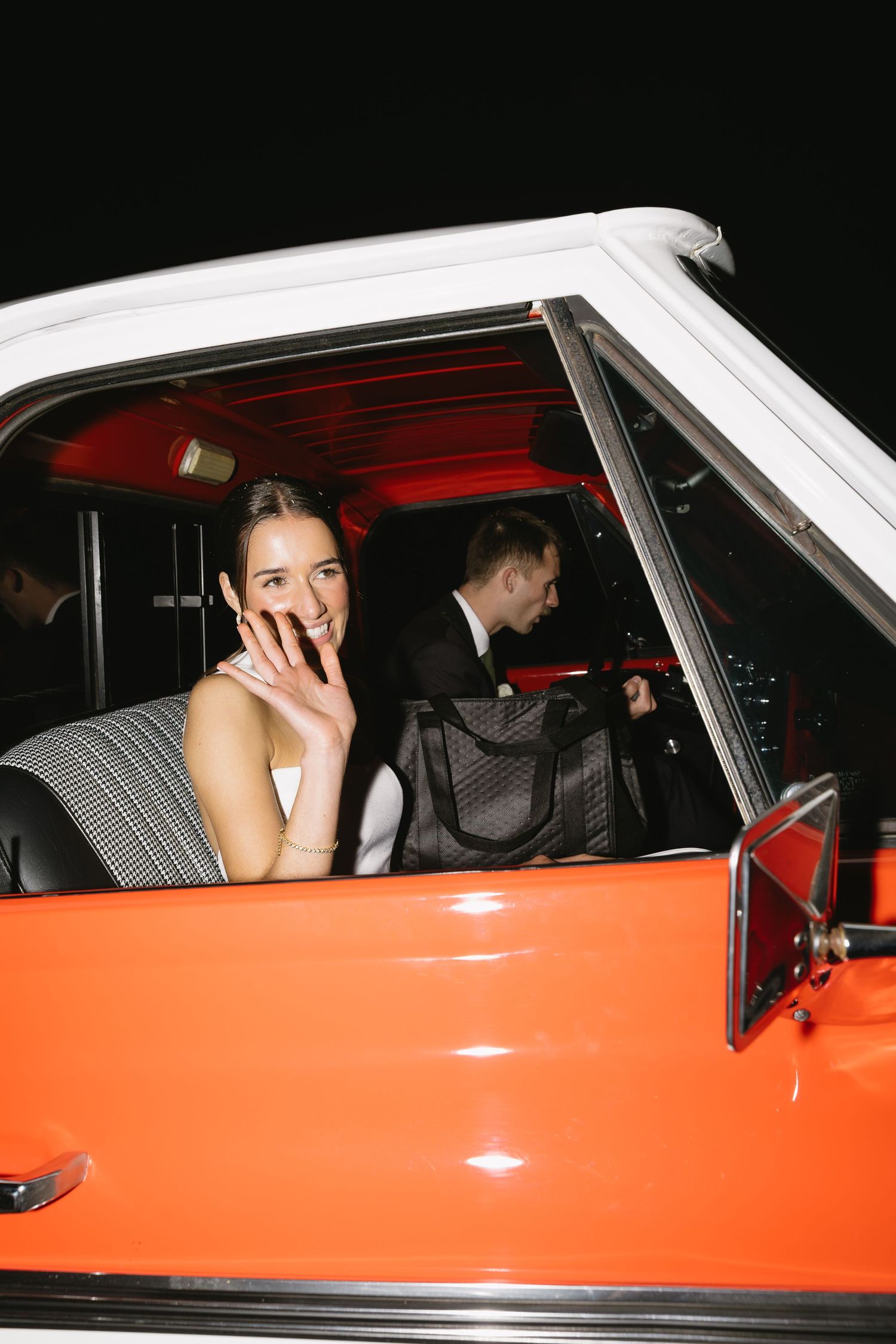 Wedding guests seated in an orange and white classic car at night.