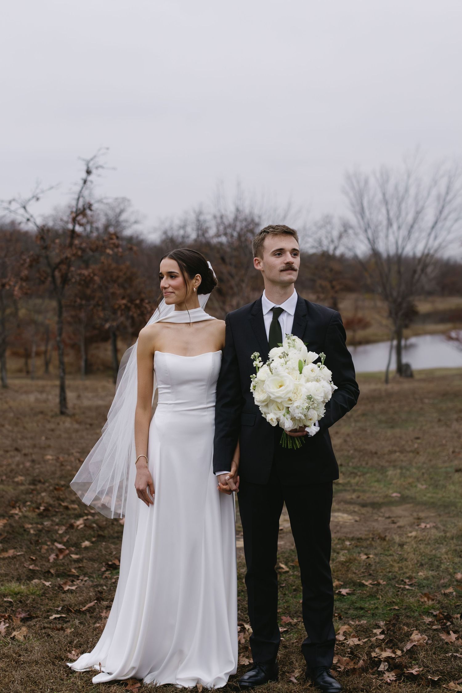 Wedding couple posing in elegant attire against rustic autumn landscape.