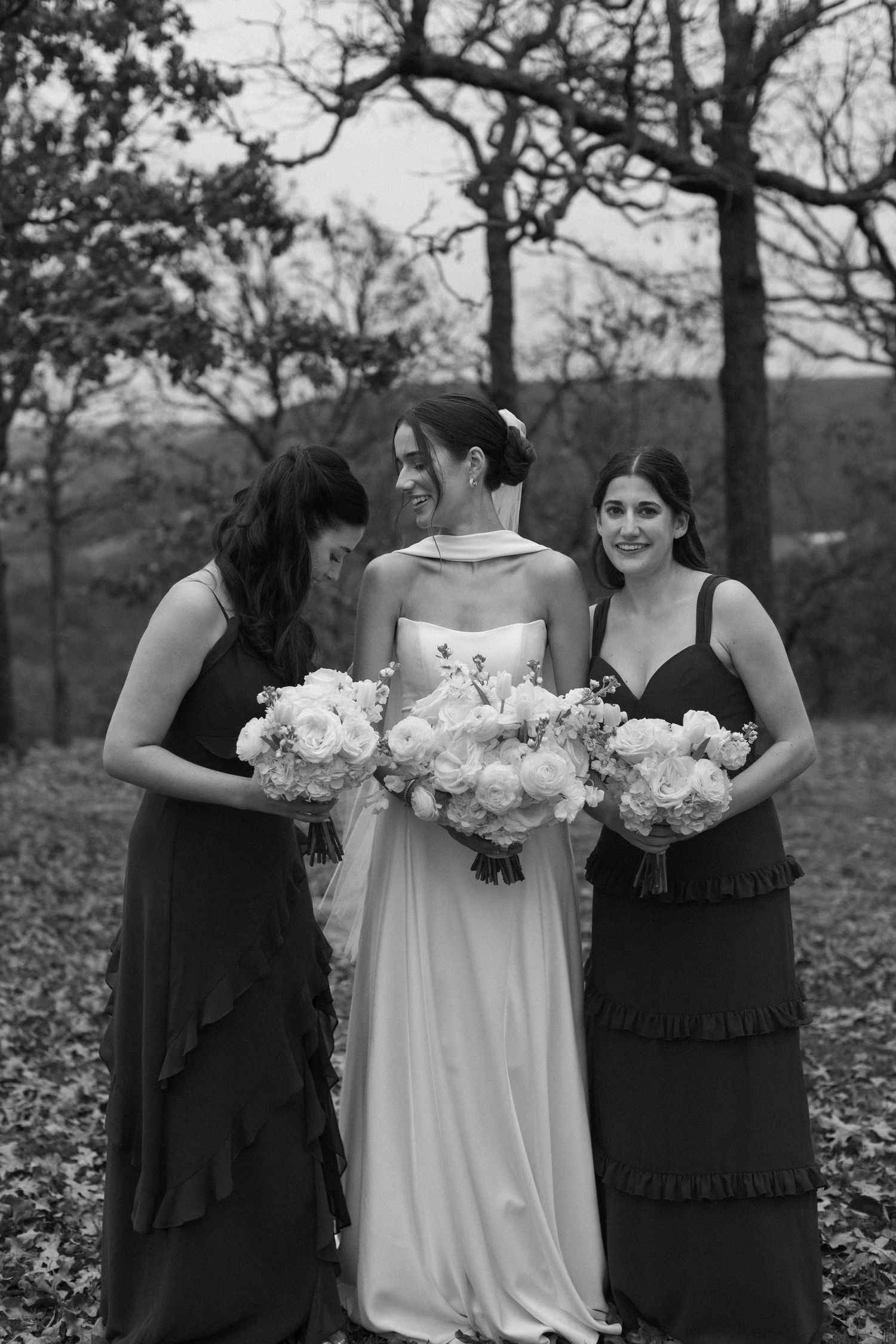 Bridal party in black dresses poses with bride in white holding bouquets outdoors.