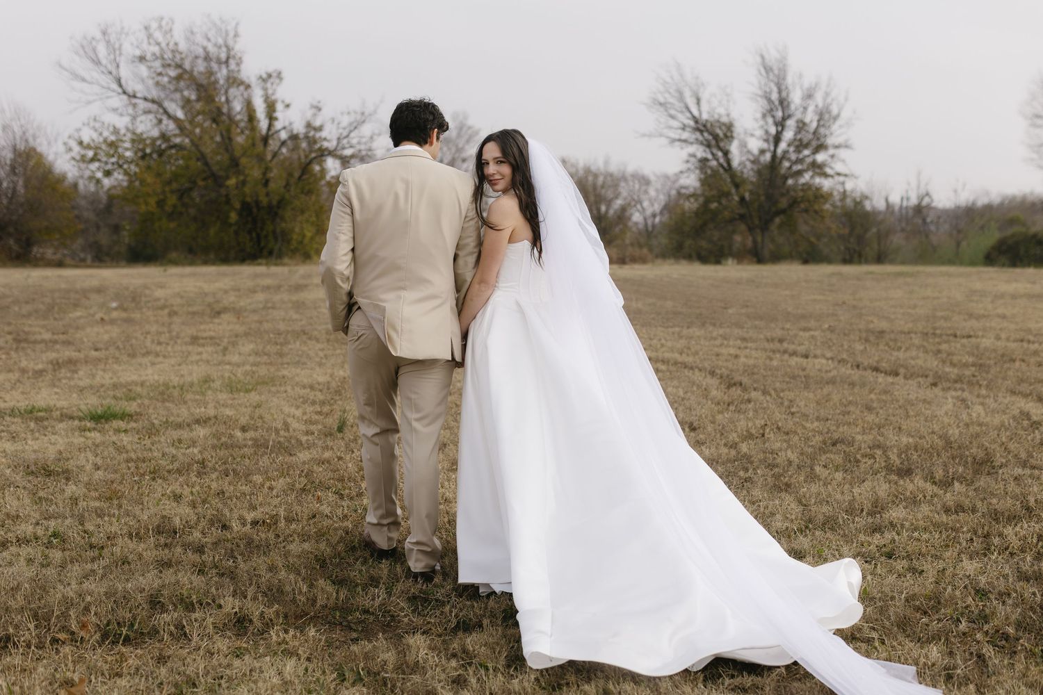 A couple in wedding attire walks hand in hand across a rustic autumn field during golden hour.