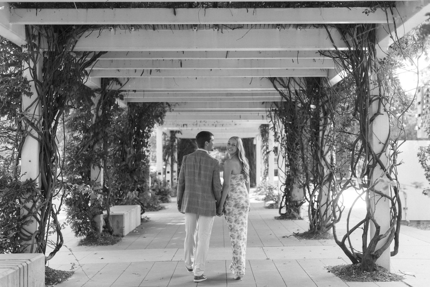 A couple walks through a pergola decorated with vines and greenery in black and white.