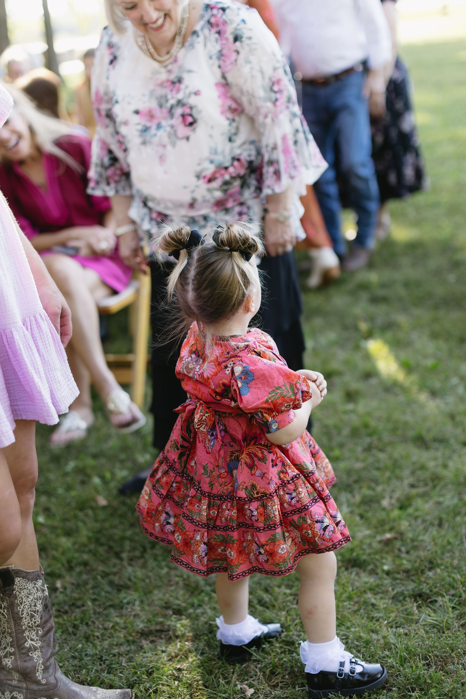 Little girl in a coral floral dress and boots dancing on the grass at a wedding celebration.