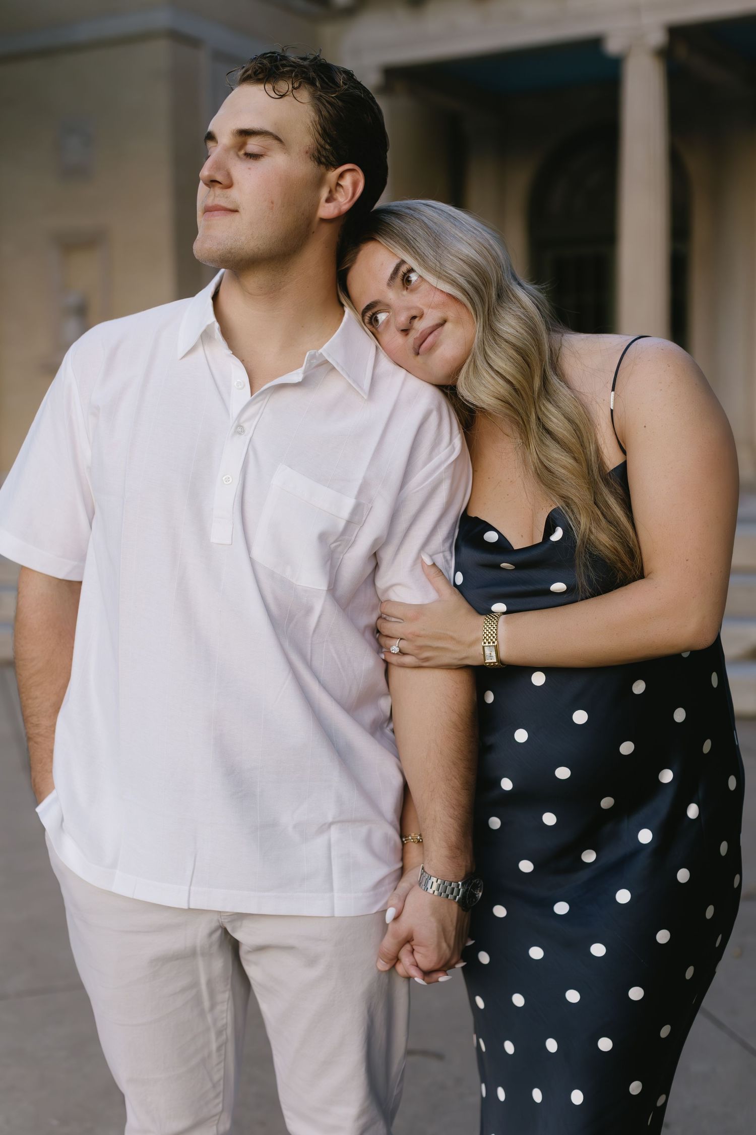 Couple leans against classical building, woman in polka dot dress rests head on man's shoulder.