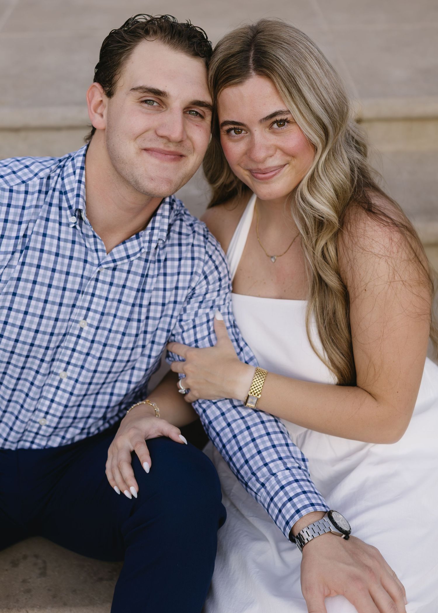A couple poses together outdoors, with one wearing a blue checkered shirt and the other in a white dress.