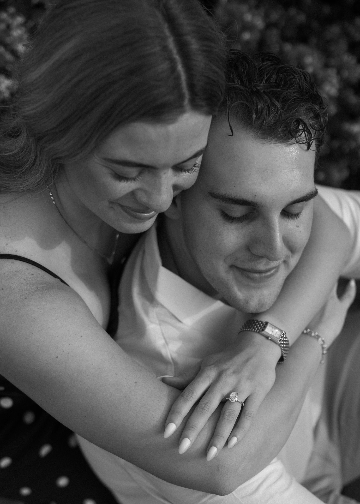 A romantic black and white portrait of a couple embracing while smiling with an engagement ring visible.