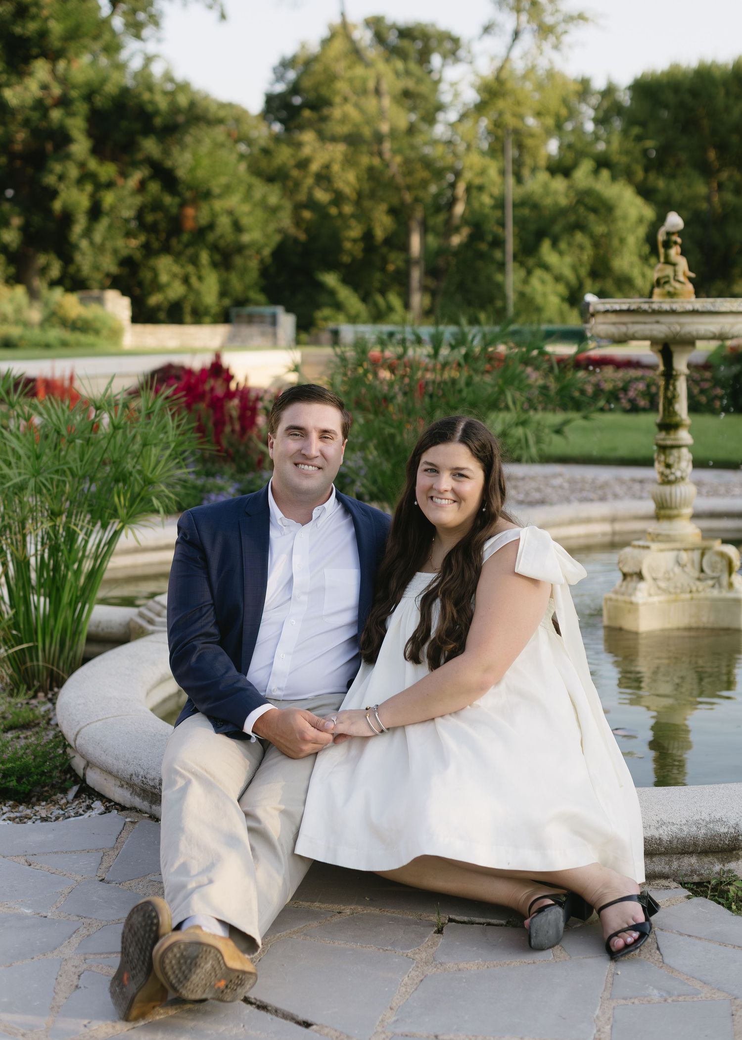 A couple poses for engagement photos near a garden fountain in formal attire.