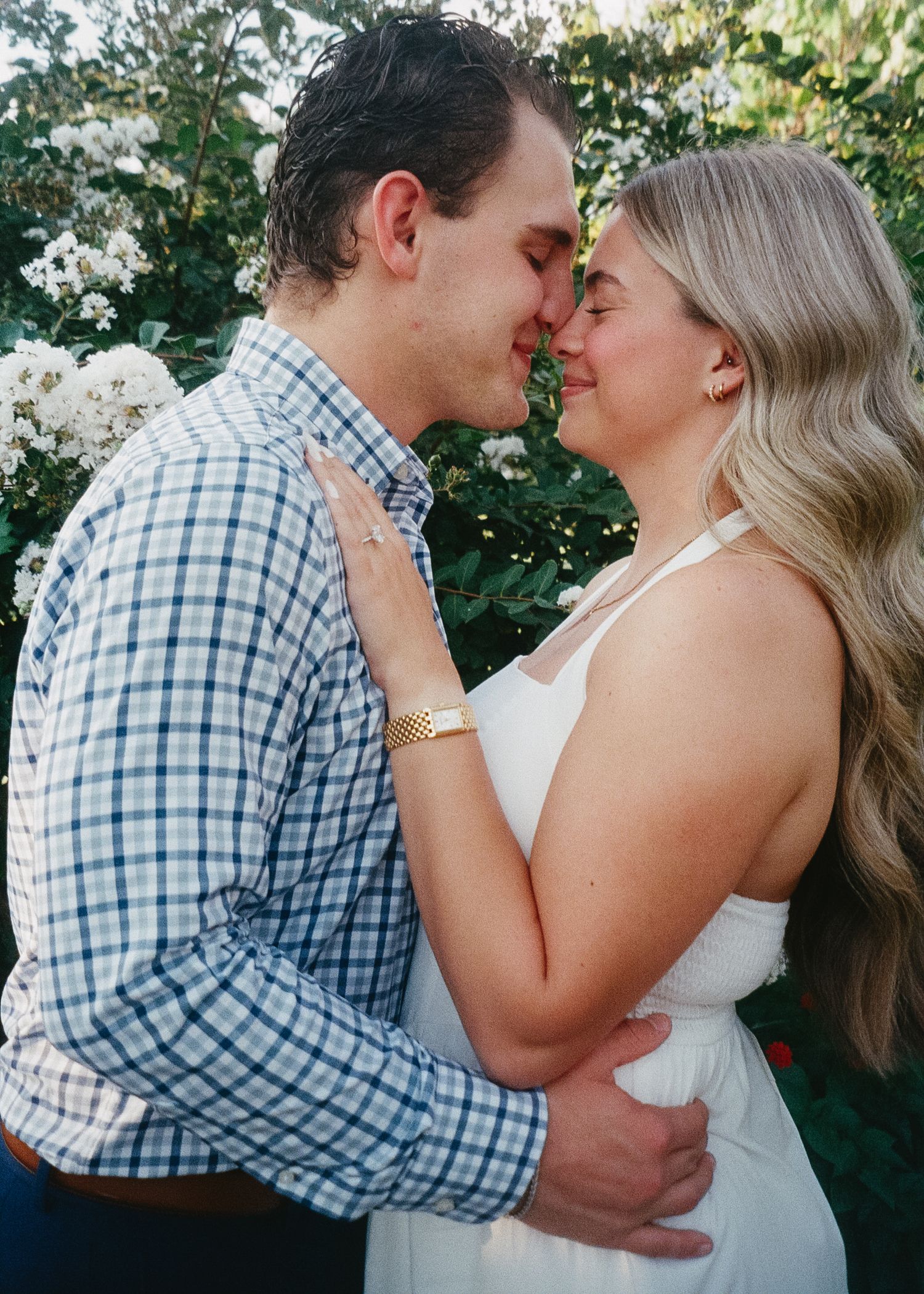 A romantic couple shares a tender kiss in a garden setting with blooming white flowers in the background.