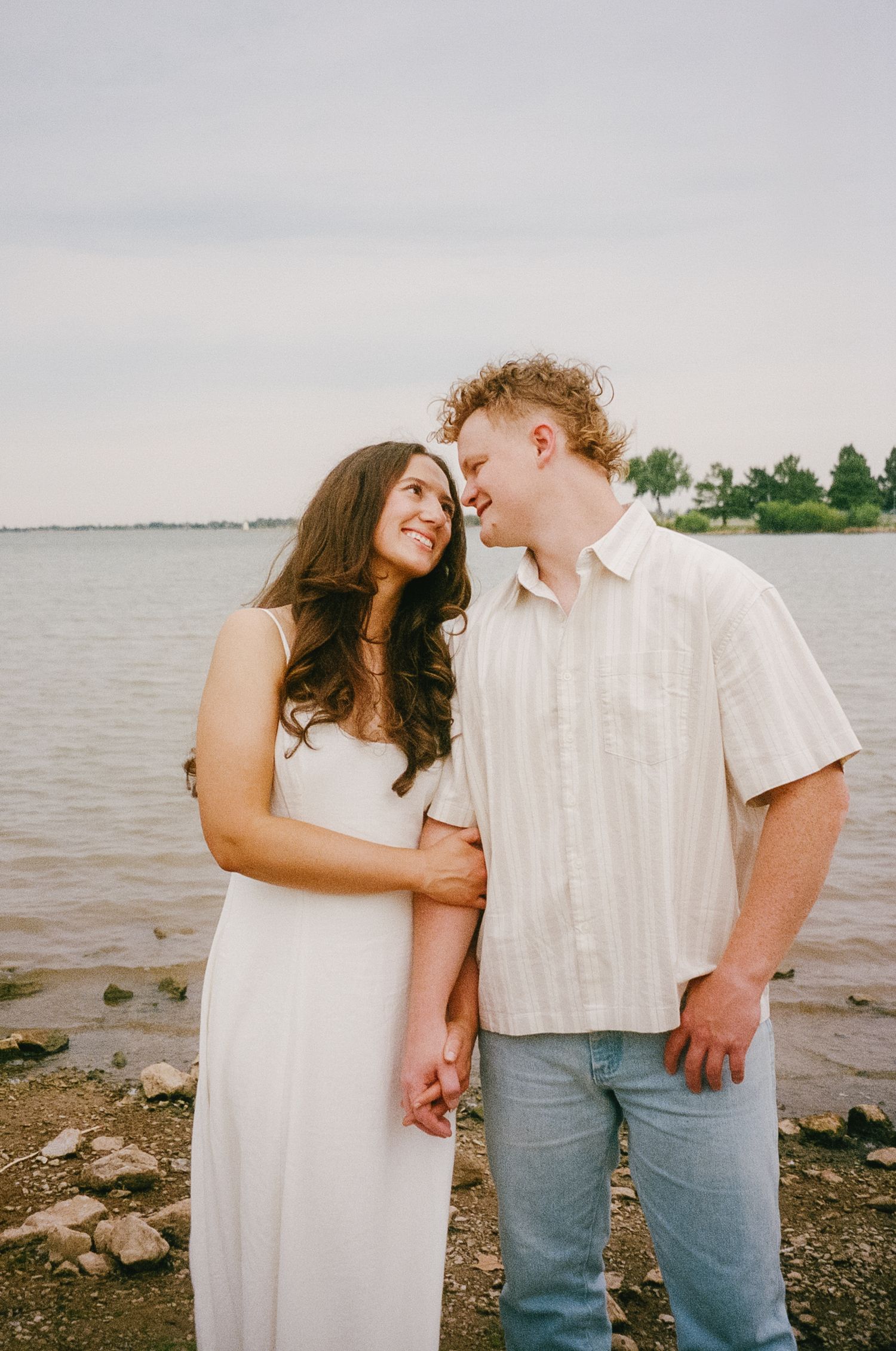 A couple in white and blue poses romantically by a lakeside on a cloudy summer day.