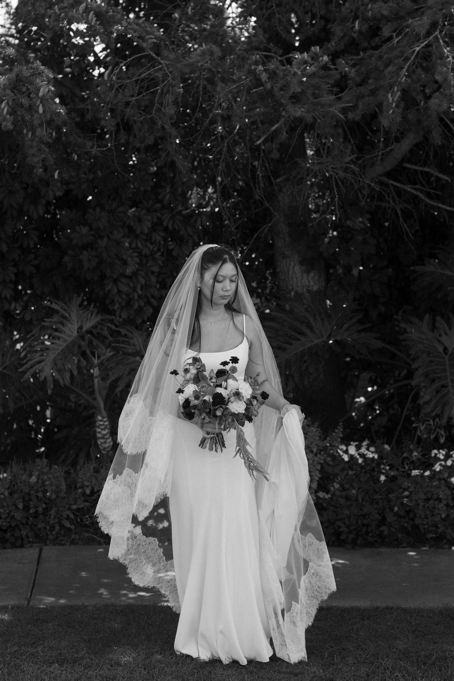 A bride in a flowing white dress and veil holds a bouquet while walking outdoors.