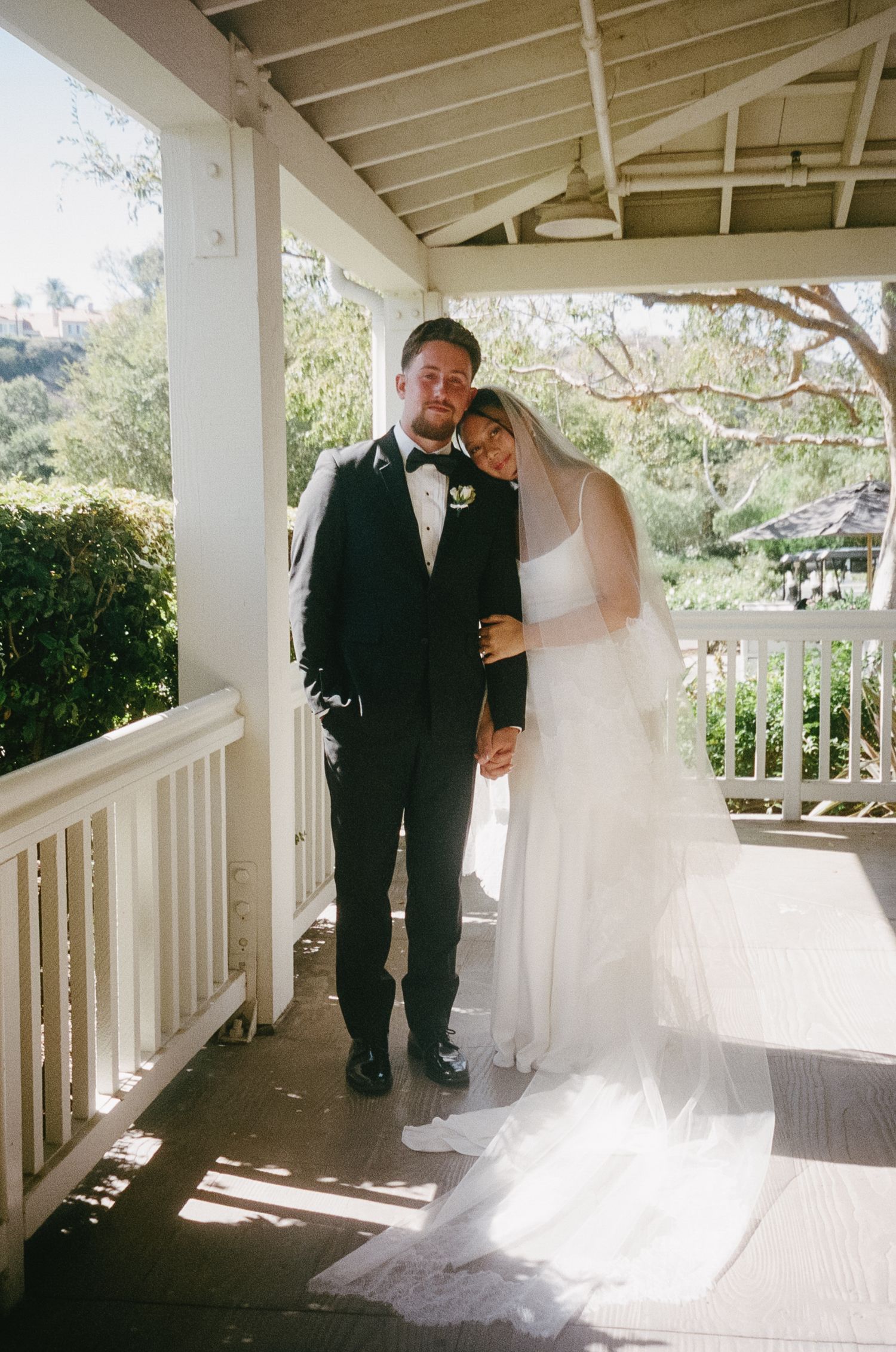 A couple in wedding attire shares intimate moments on a sunlit white porch.