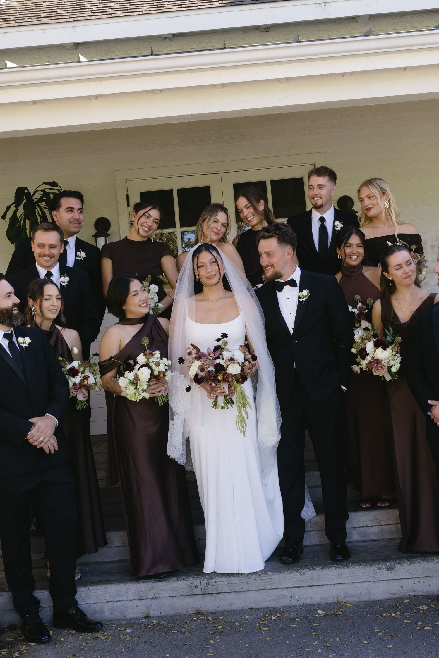 Wedding party in formal black and brown attire poses together on steps of white building during outdoor ceremony.