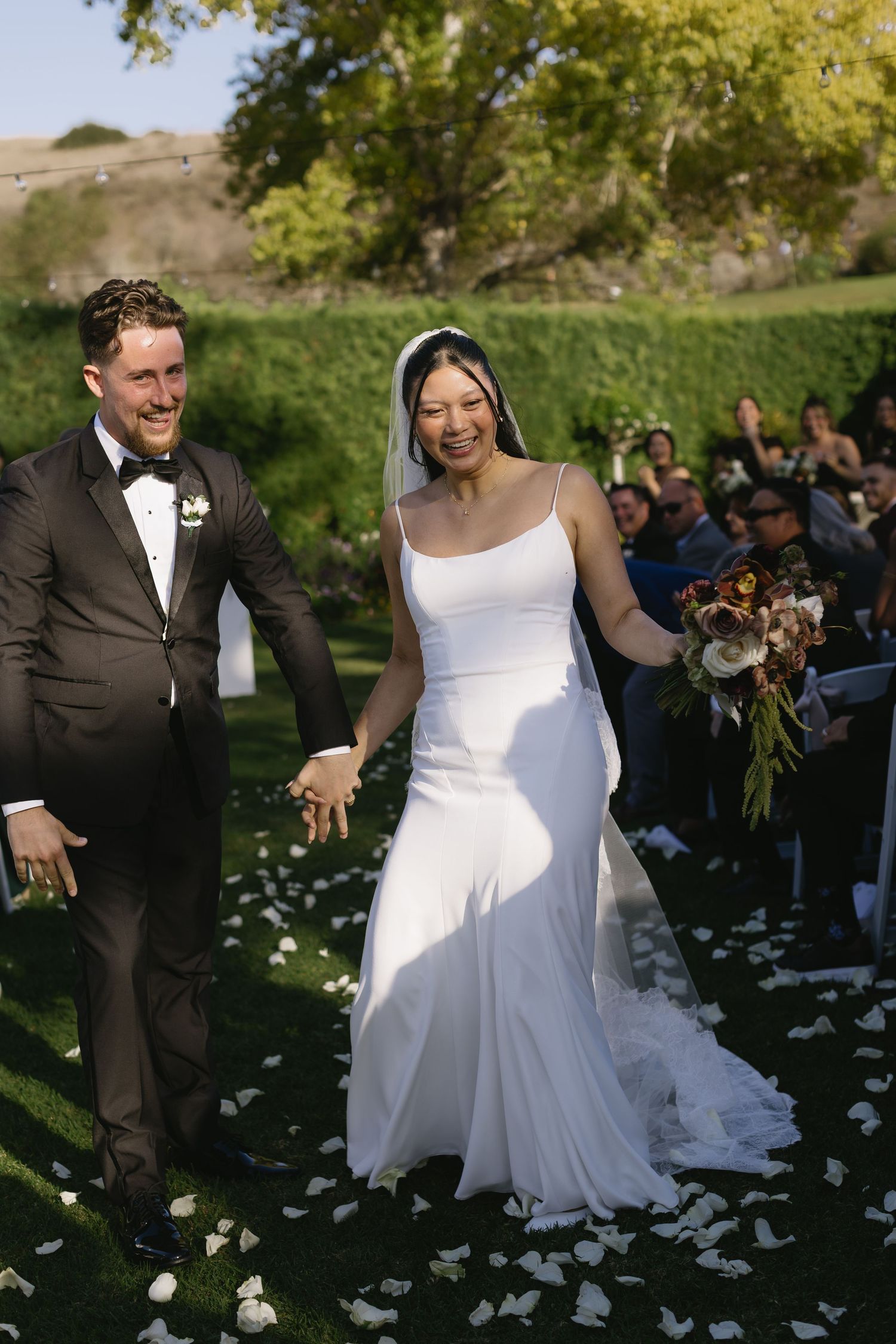 Newlyweds walk down aisle strewn with white rose petals in outdoor garden ceremony.
