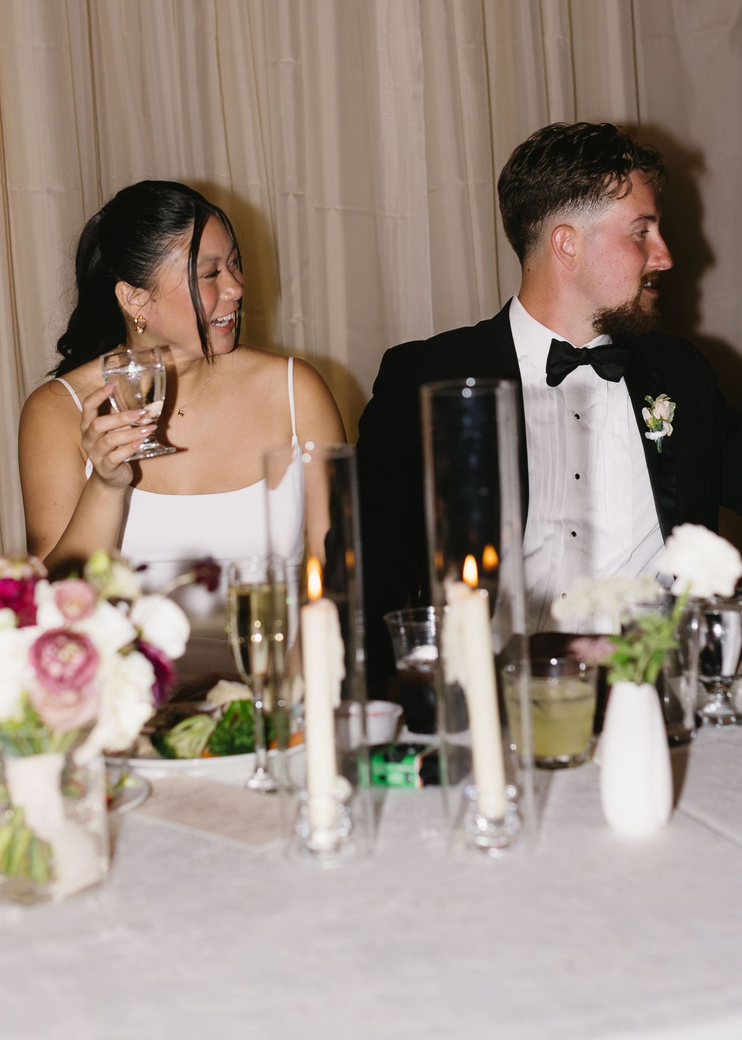 A couple shares laughter at their wedding reception head table surrounded by elegant decor.
