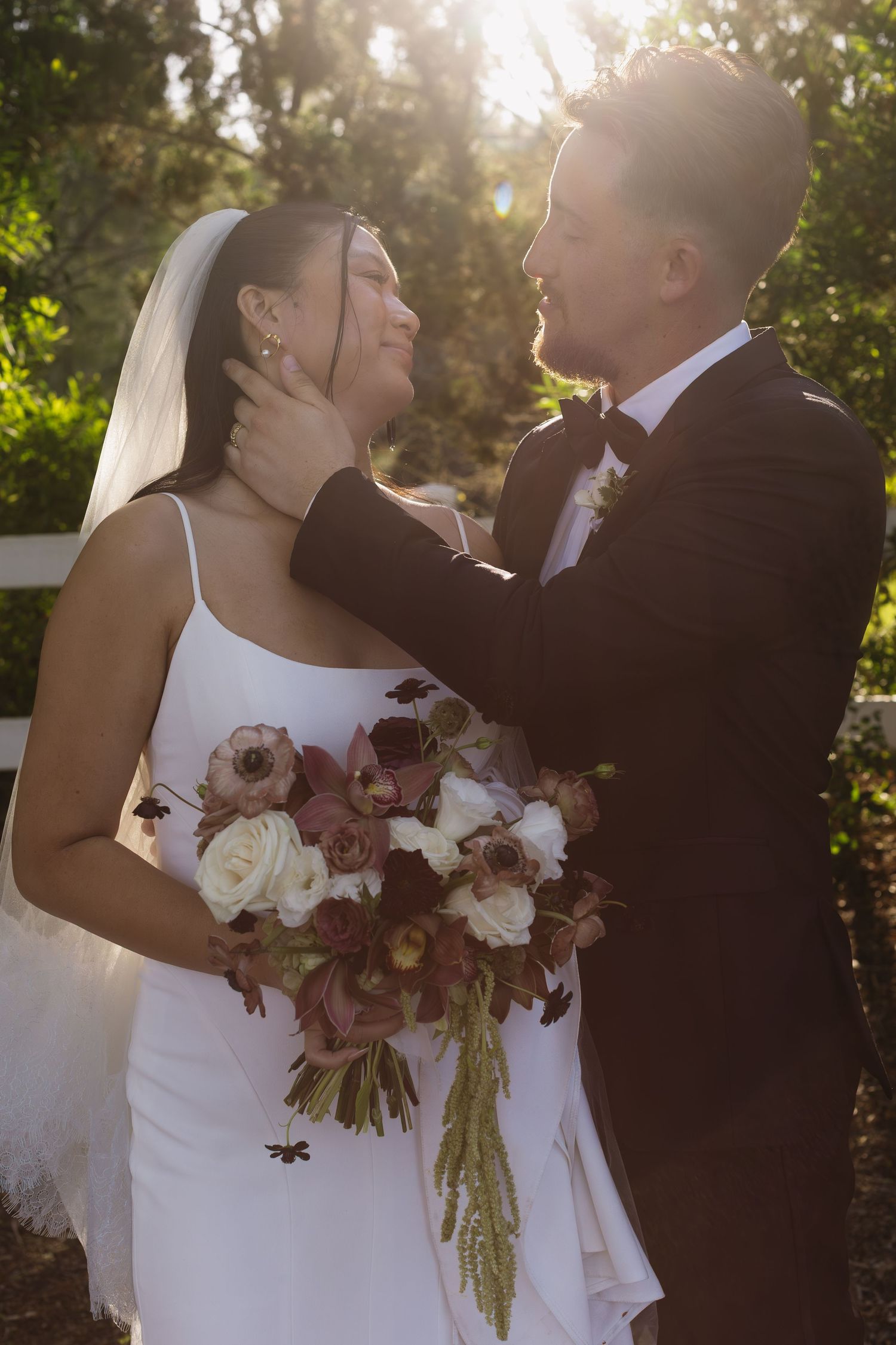Newlyweds share an intimate moment in golden sunlight with burgundy and white floral bouquet.