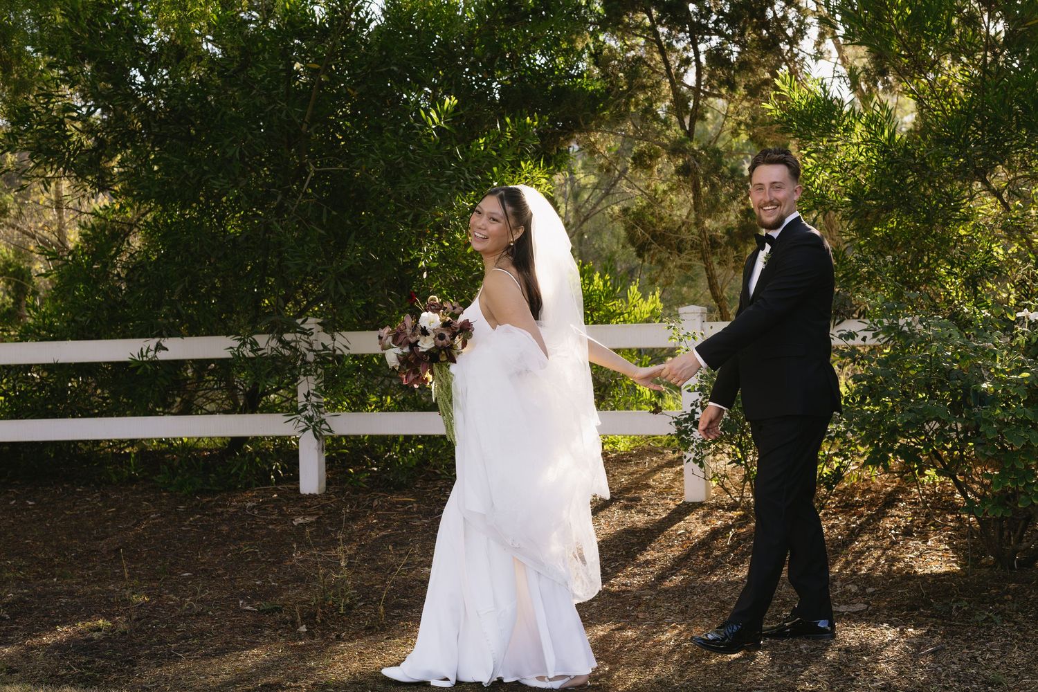 A wedding couple poses together in a lush garden setting with trees and greenery in the background.