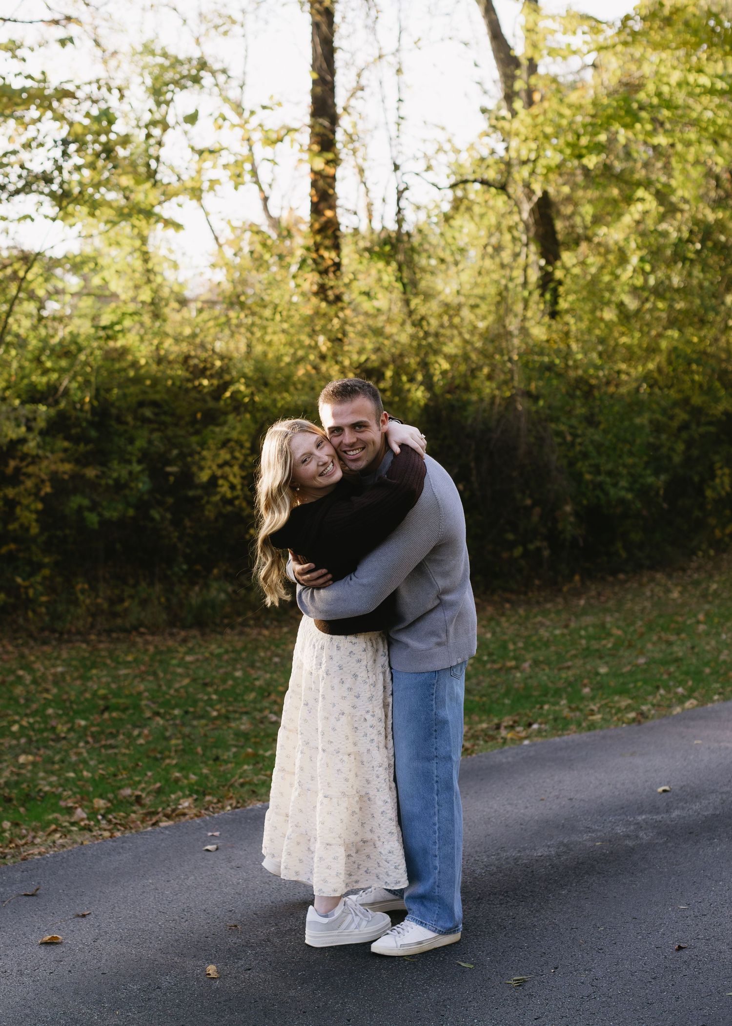 A couple shares a playful moment on a paved path surrounded by autumn foliage.