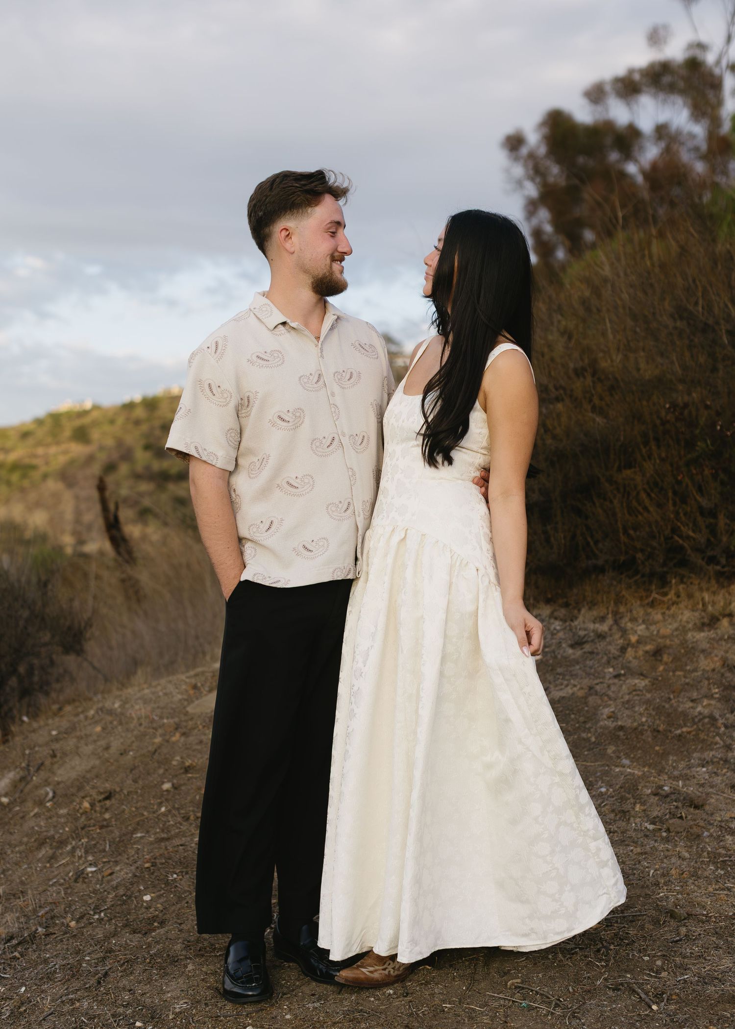 A couple in coordinated white outfits poses together on a hillside trail during a romantic photoshoot.