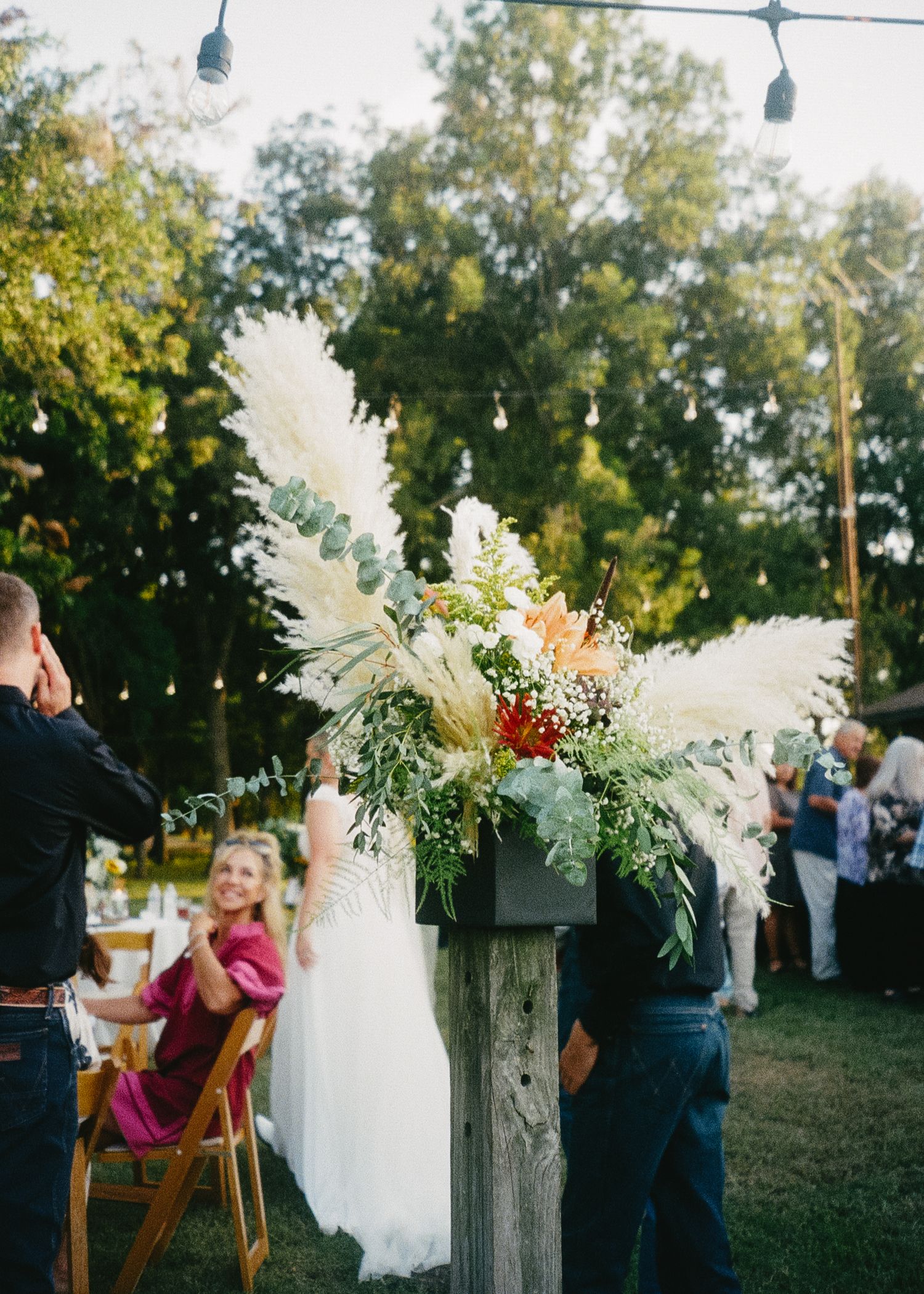 Outdoor wedding reception setup with string lights, white floral arrangements, and wooden chairs at dusk.