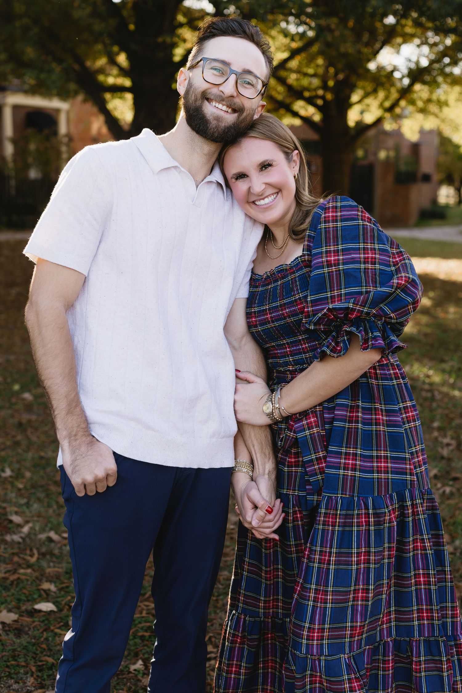 A couple stands together outdoors, with one wearing a white polo and the other in a plaid dress.