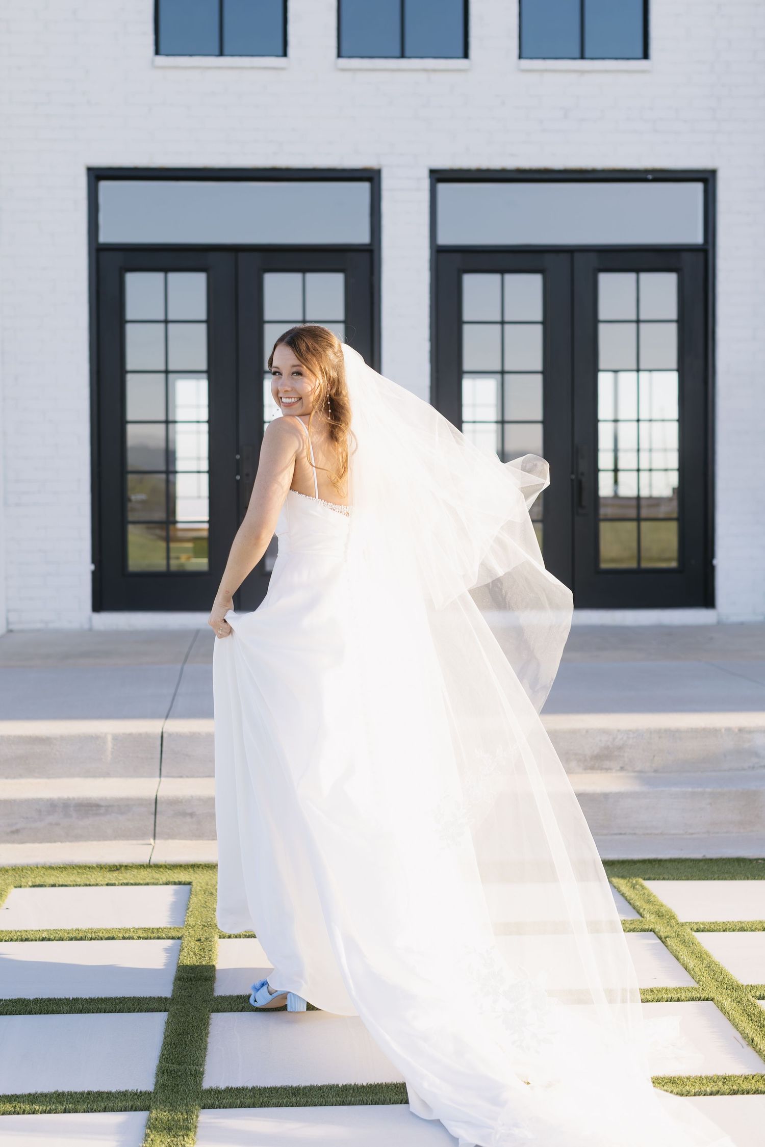 A flowing white wedding dress and veil billowing in the wind against a modern white building with black windows.