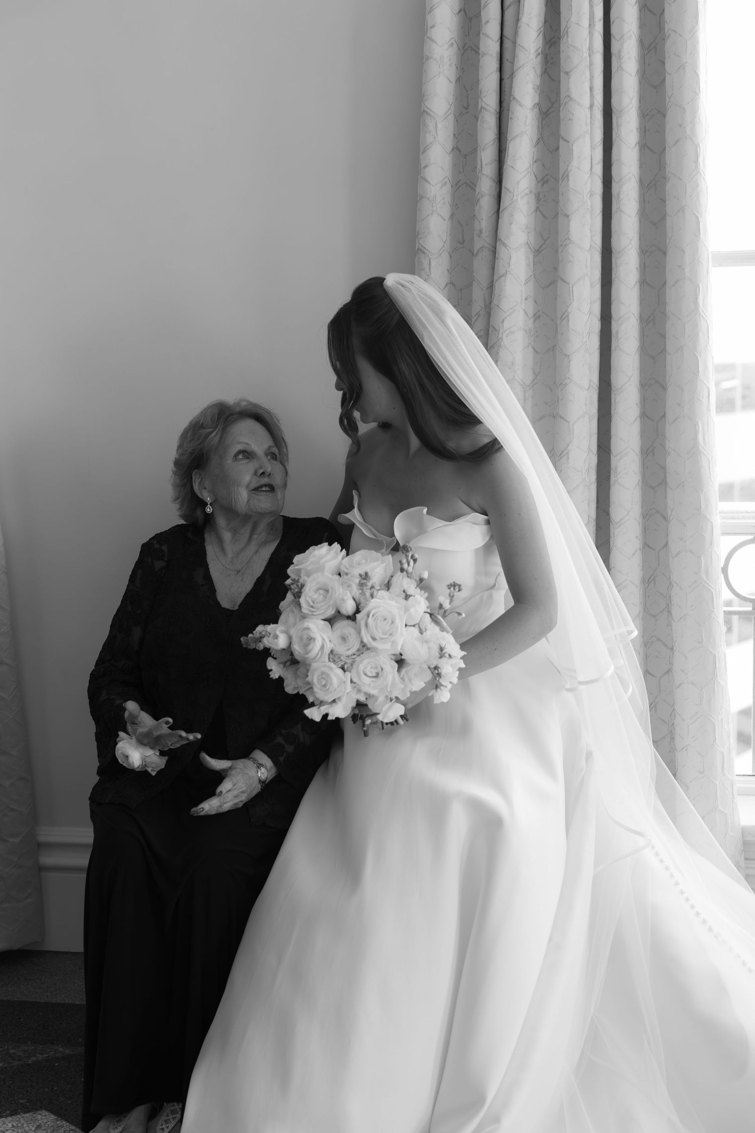 Black and white wedding photo showing an elder woman in dark dress seated with bride in white gown holding rose bouquet.