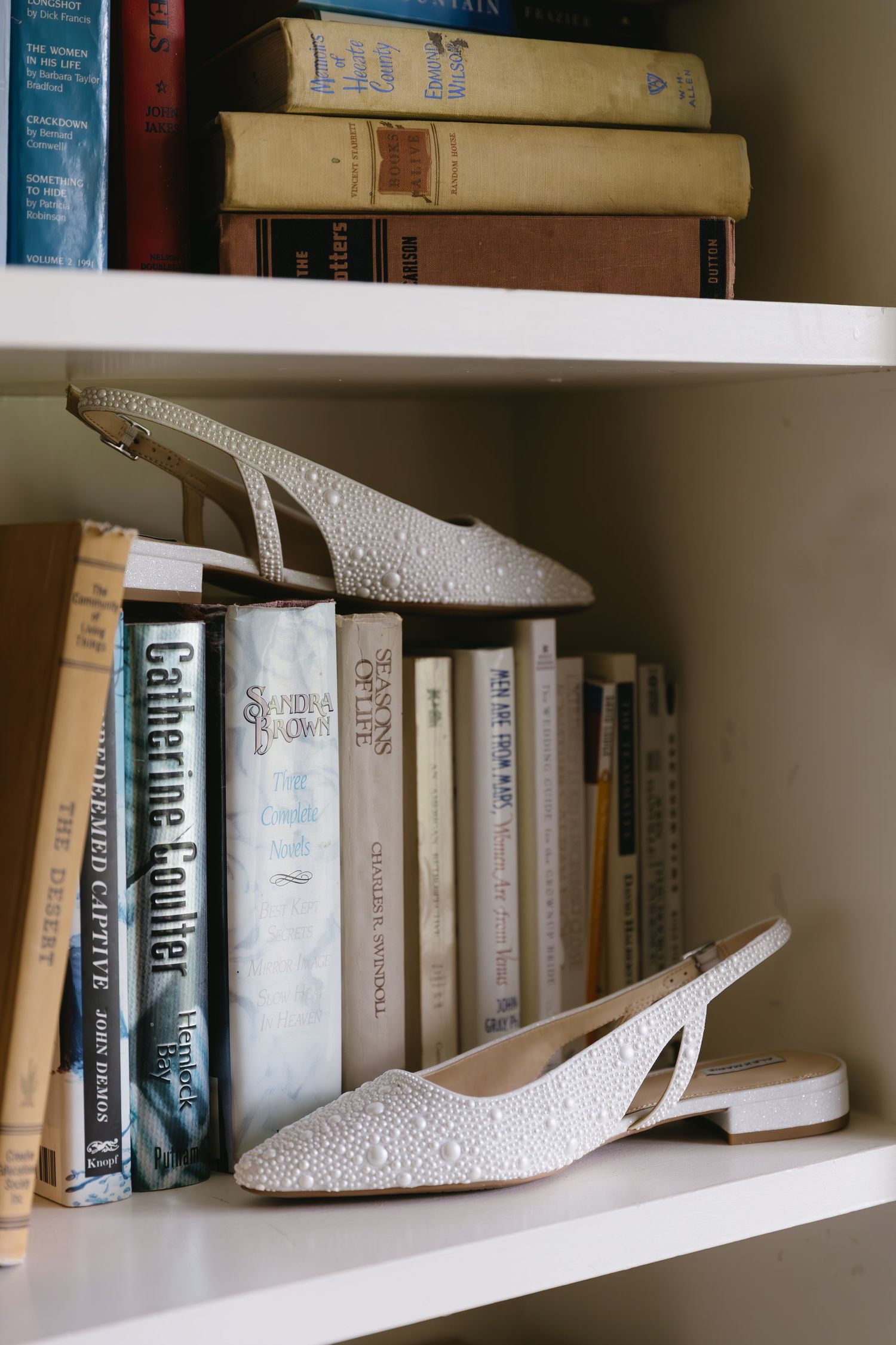 White lacey slingback shoes displayed on wooden bookshelf between rows of books.