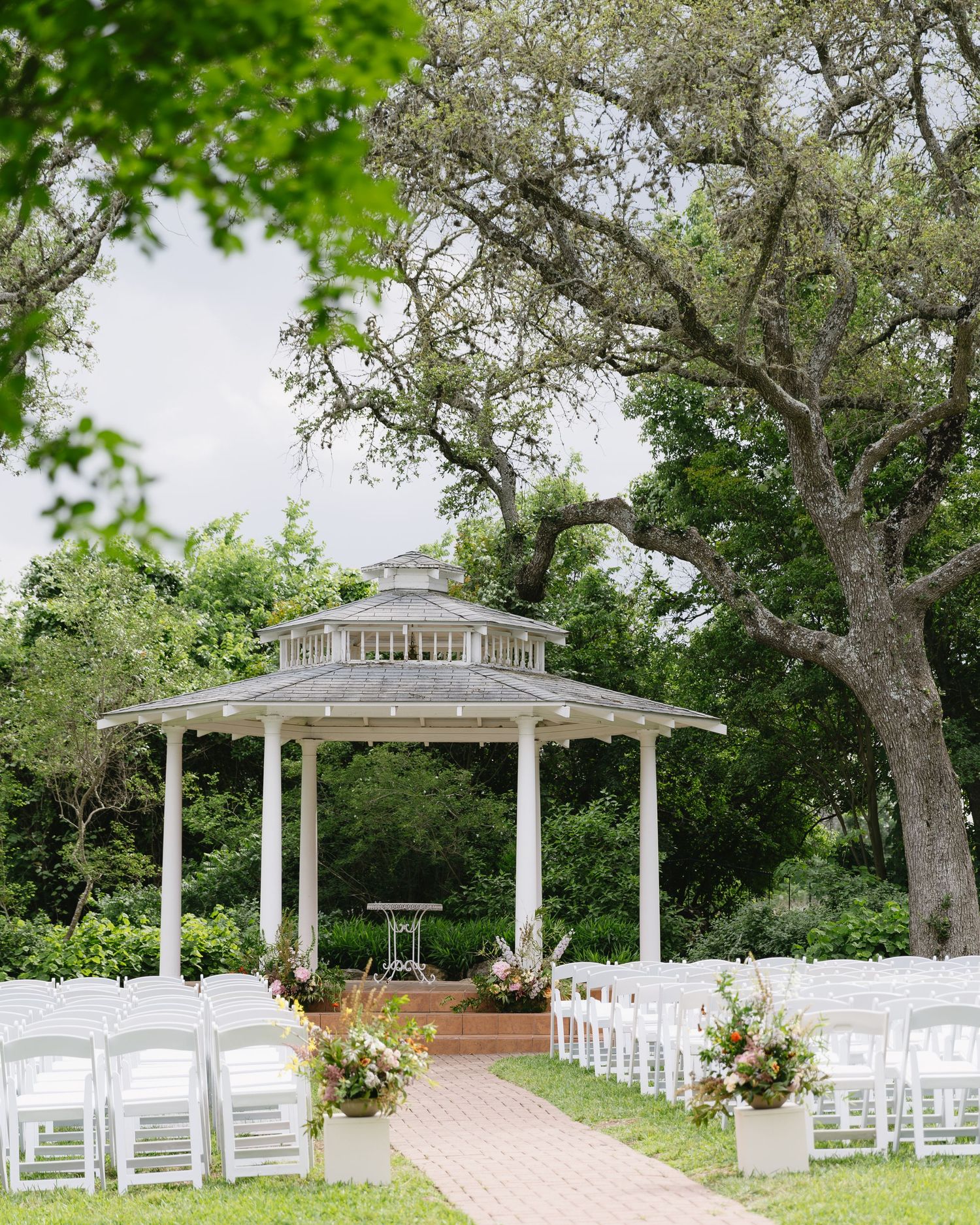 Elegant outdoor wedding ceremony setup with white chairs and gazebo under large oak trees.