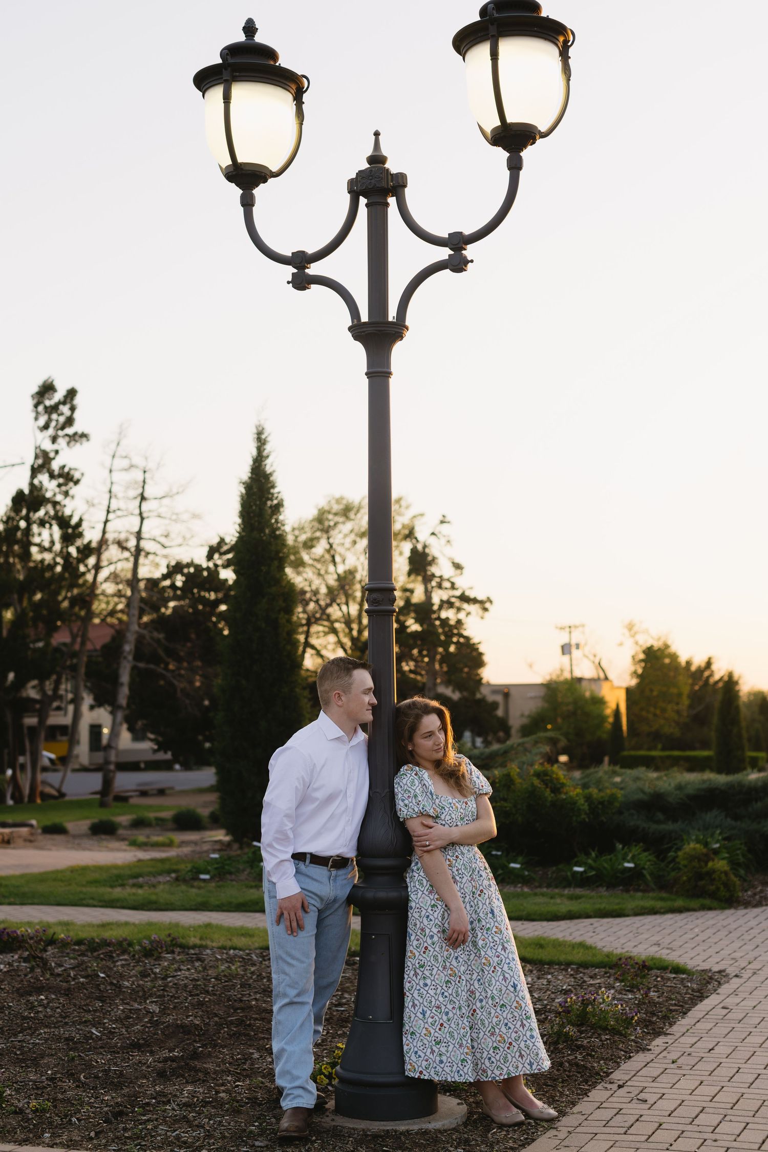 A couple poses beneath vintage street lamps along a brick pathway at dusk.