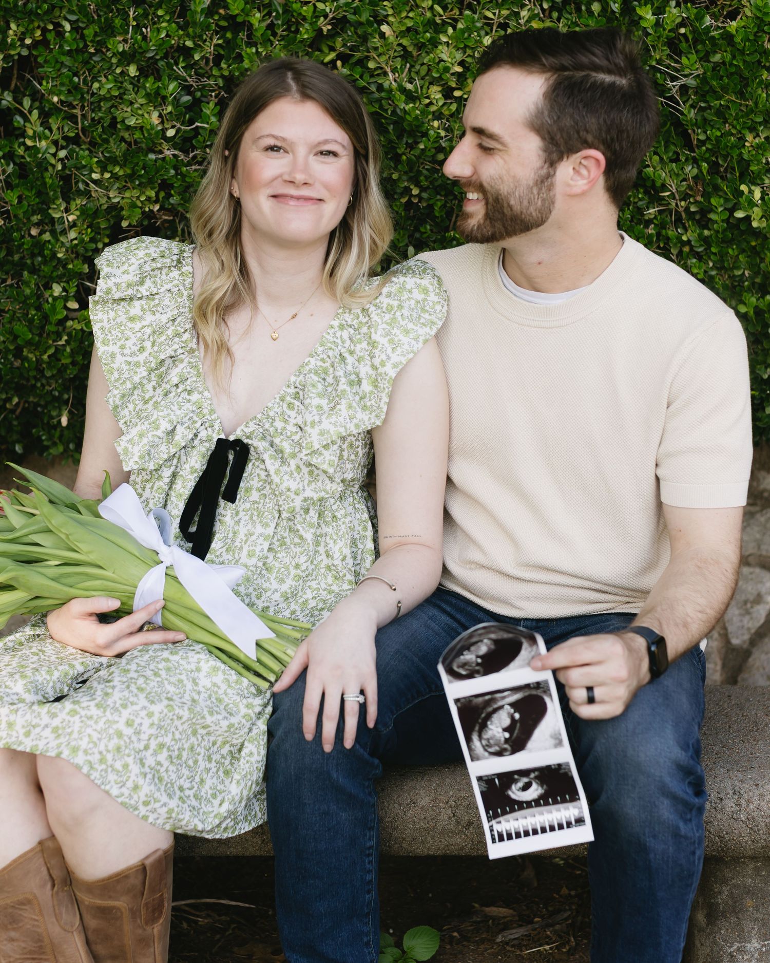 Couple sitting outdoors holding sonogram photos and sharing a tender moment among green foliage.