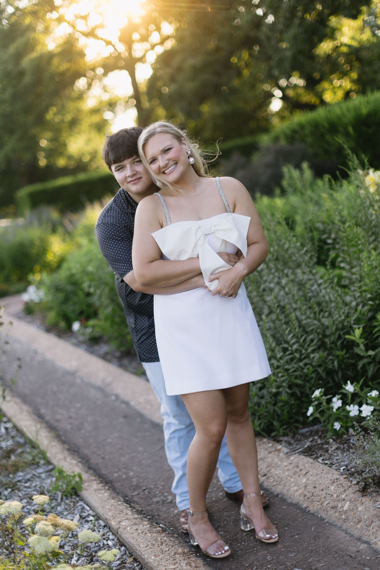 Playful couple posing together on a garden path during golden hour sunset with greenery in background.