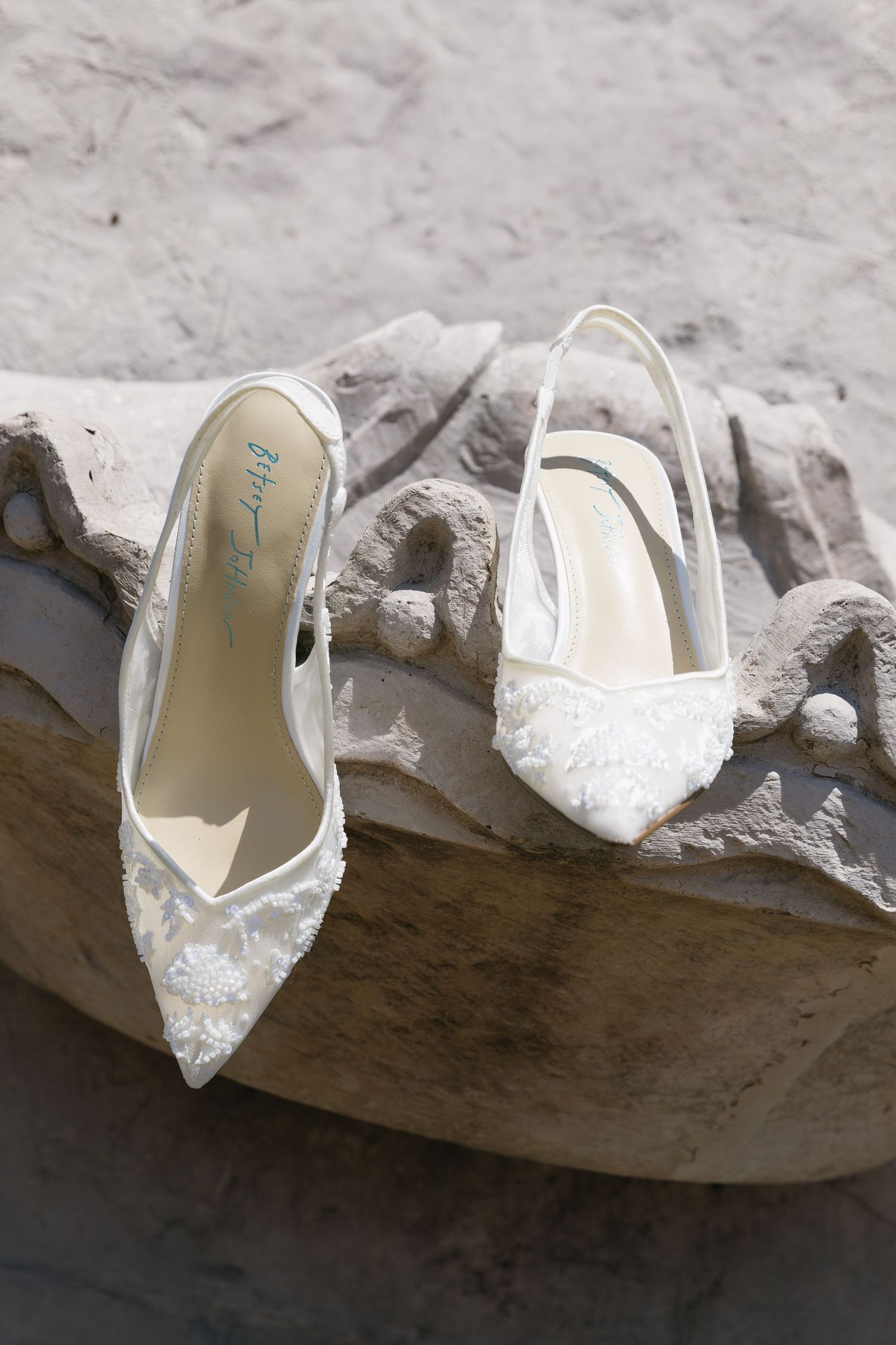 Delicate white lace wedding shoes with intricate beading displayed on a stone surface.