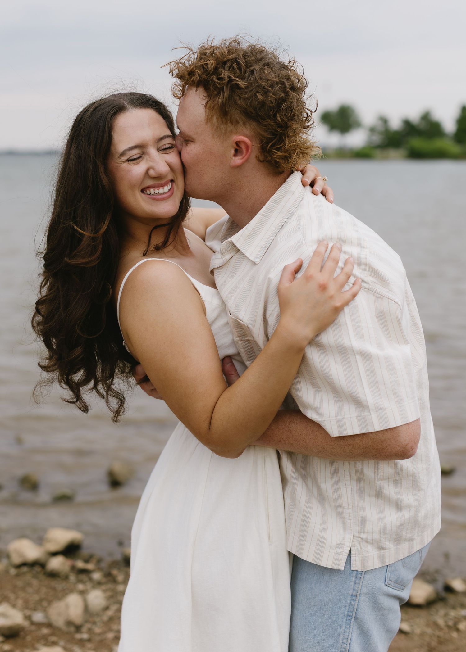 Couple shares intimate moment near rocky waterfront wearing casual white and denim outfits.